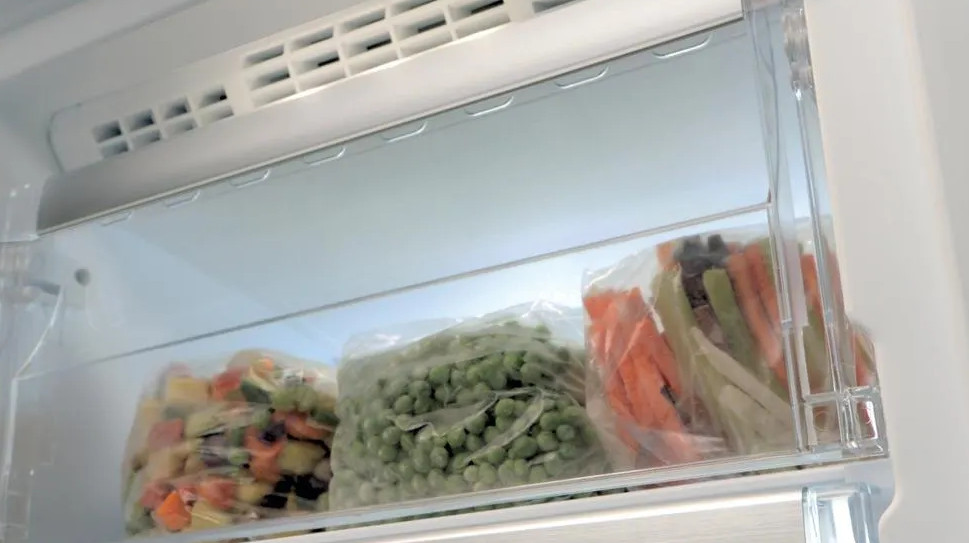 Wewnątrz zamrażarki trzy worki mrożonych warzyw: mieszanka, groszek i marchew z selerem.