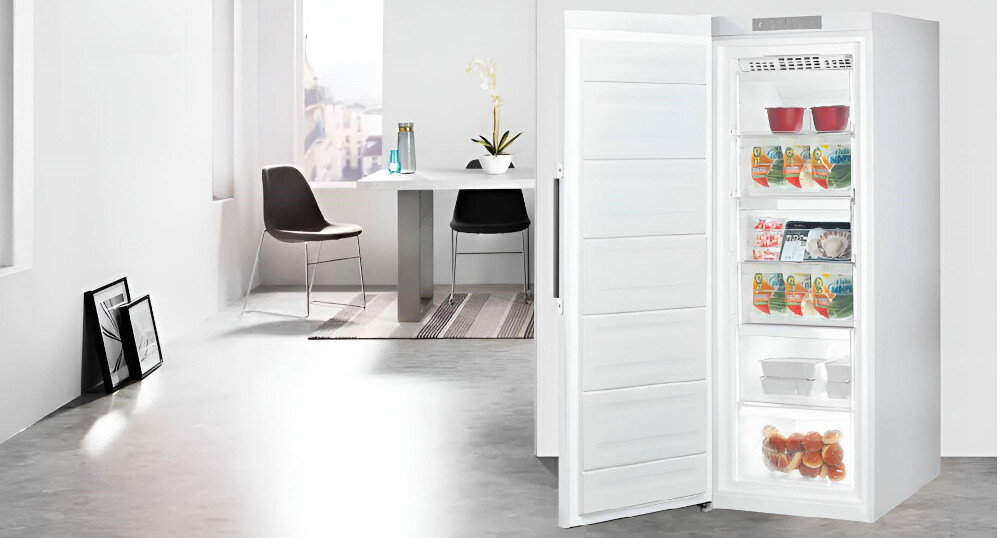 Biała zamrażarka z otwartymi drzwiami, pokazująca szuflady z jedzeniem. W tle stół jadalny z krzesłami i oprawionymi obrazami.