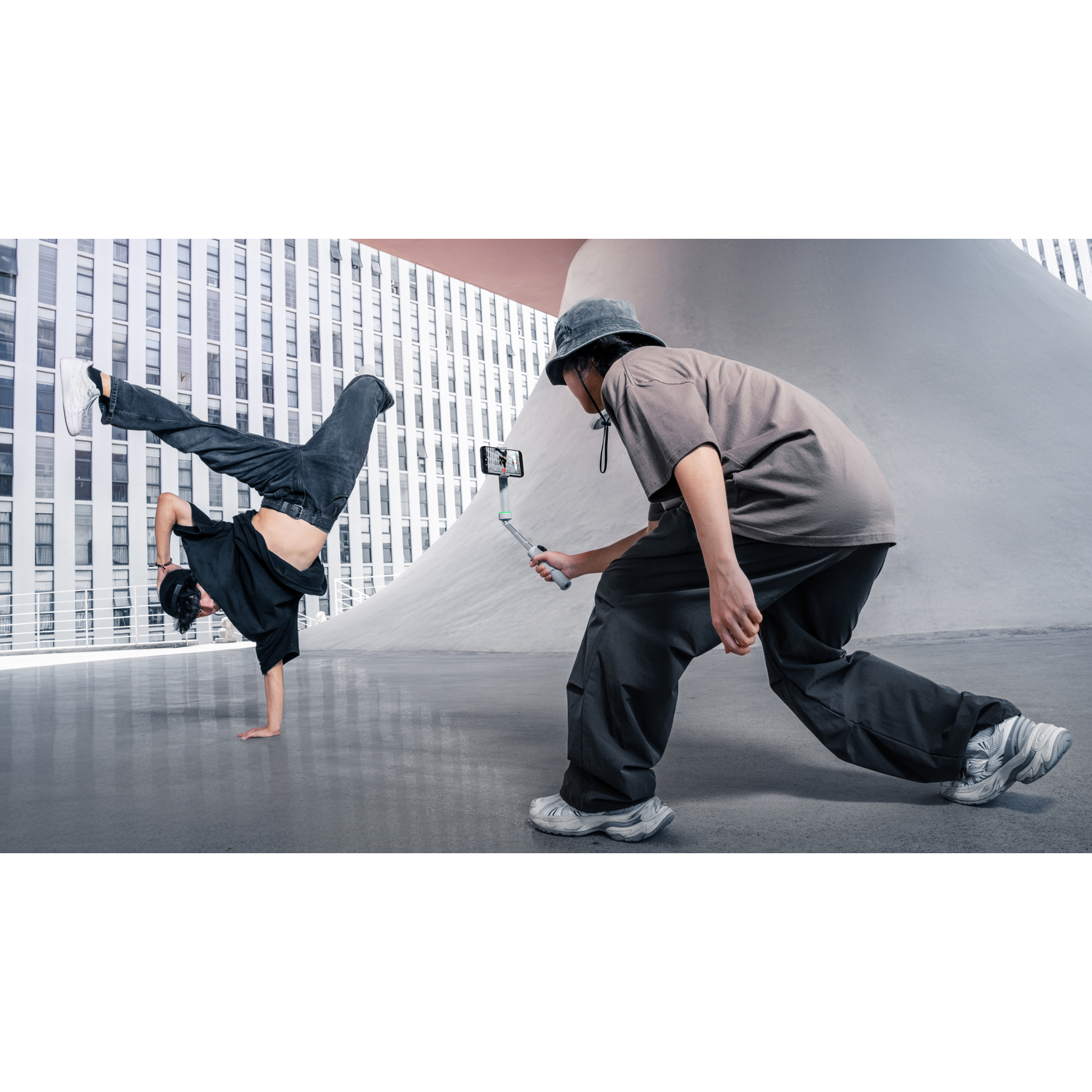 Twee mensen breakdancen buiten. Eén doet een handstand, de ander filmt met een telefoon. Moderne architectuur op de achtergrond.