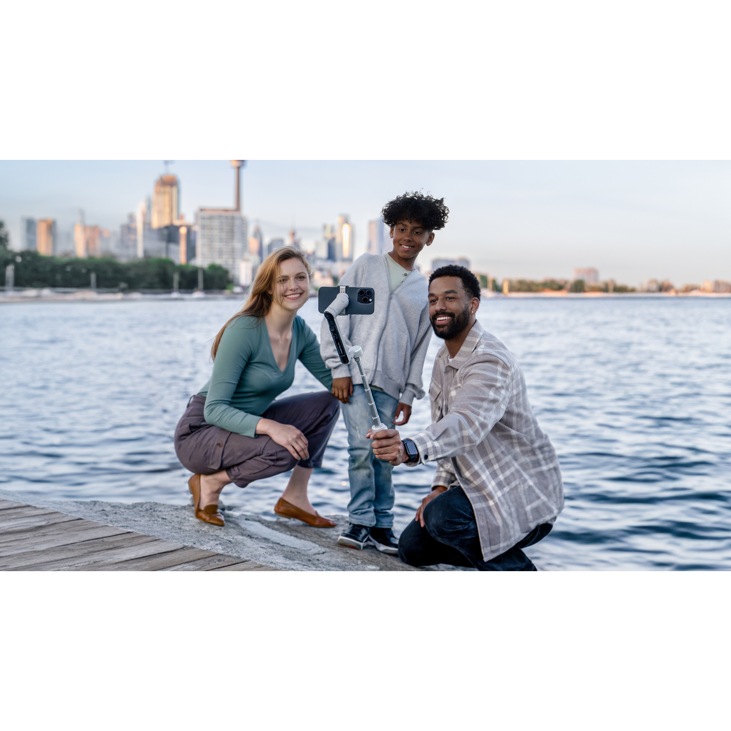 Een gezin van drie maakt een selfie bij het water. Een skyline is op de achtergrond. Ze gebruiken een selfiestick.