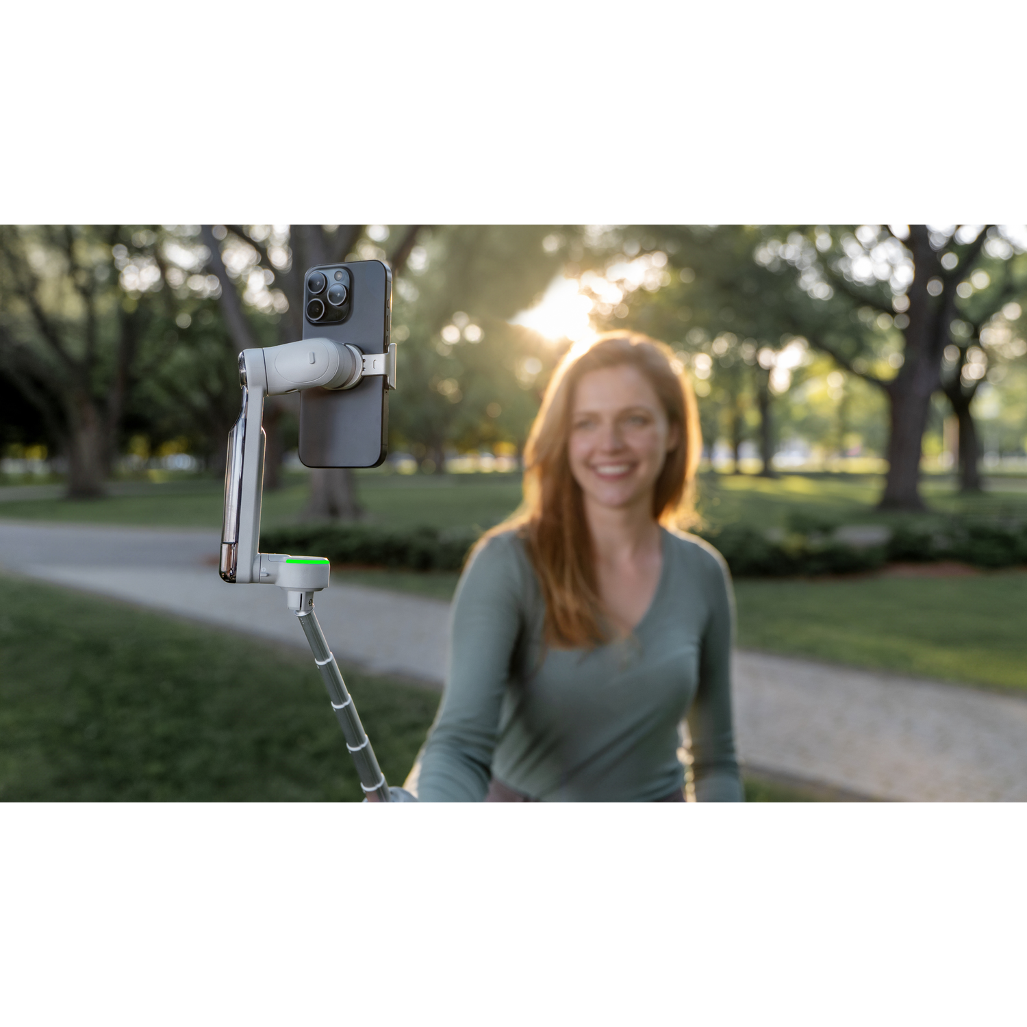 Een vrouw glimlacht, filmt met een telefoon op een selfiestick in een park. De zon schijnt door de bomen op de achtergrond.