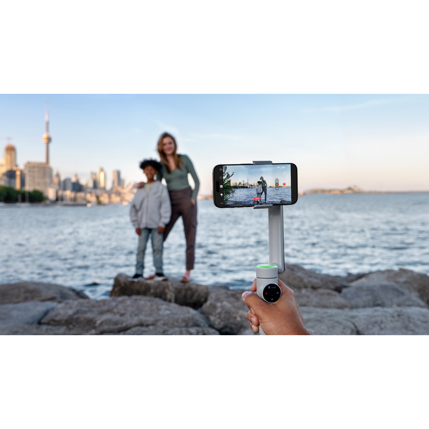 Een persoon houdt een telefoon op een gimbal. De telefoon toont twee personen. De achtergrond bevat een skyline en het meer.