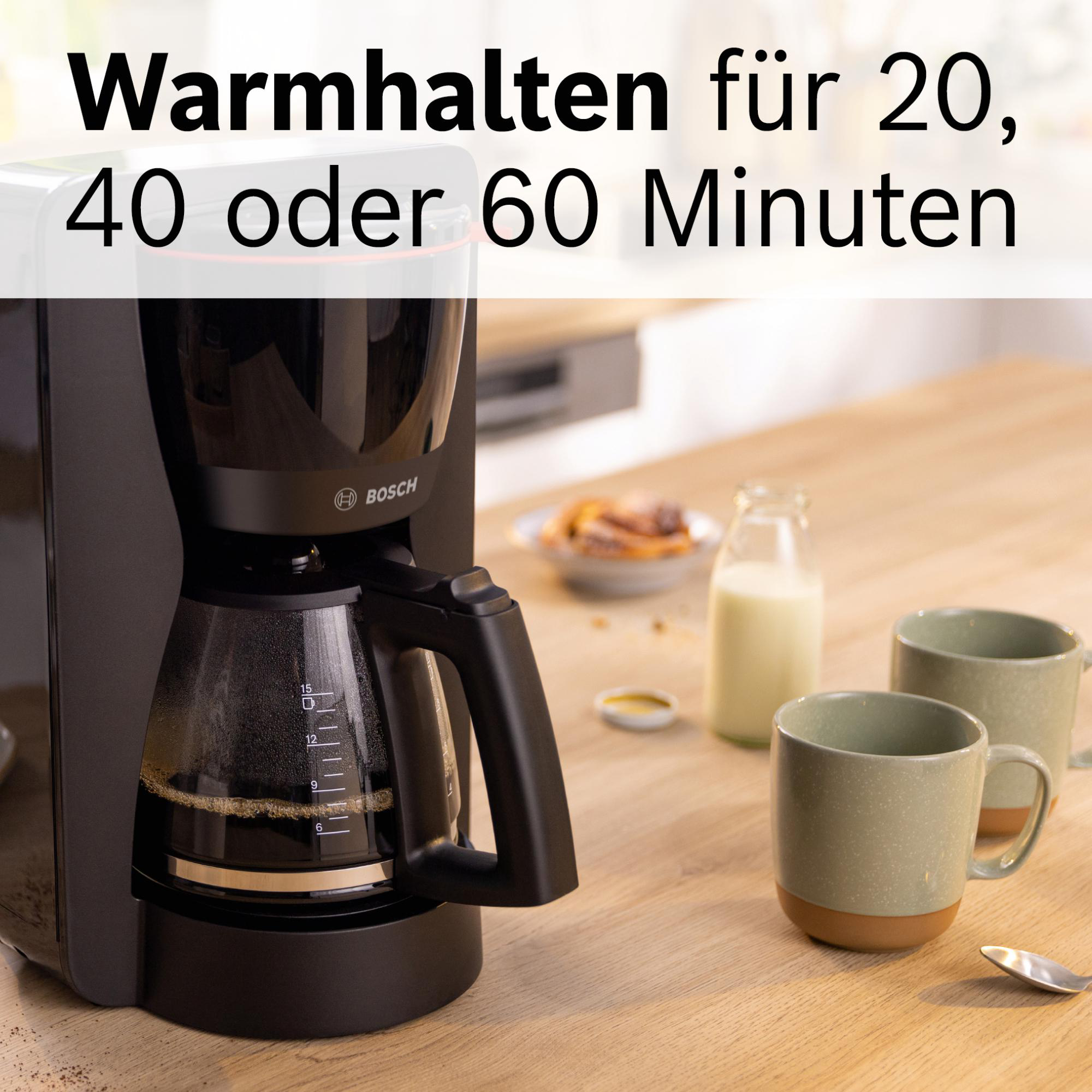 Eine schwarze Kaffeemaschine mit Glaskaraffe auf einem Holztisch. Zwei Tassen, Milch und Gebäck sind ebenfalls vorhanden.