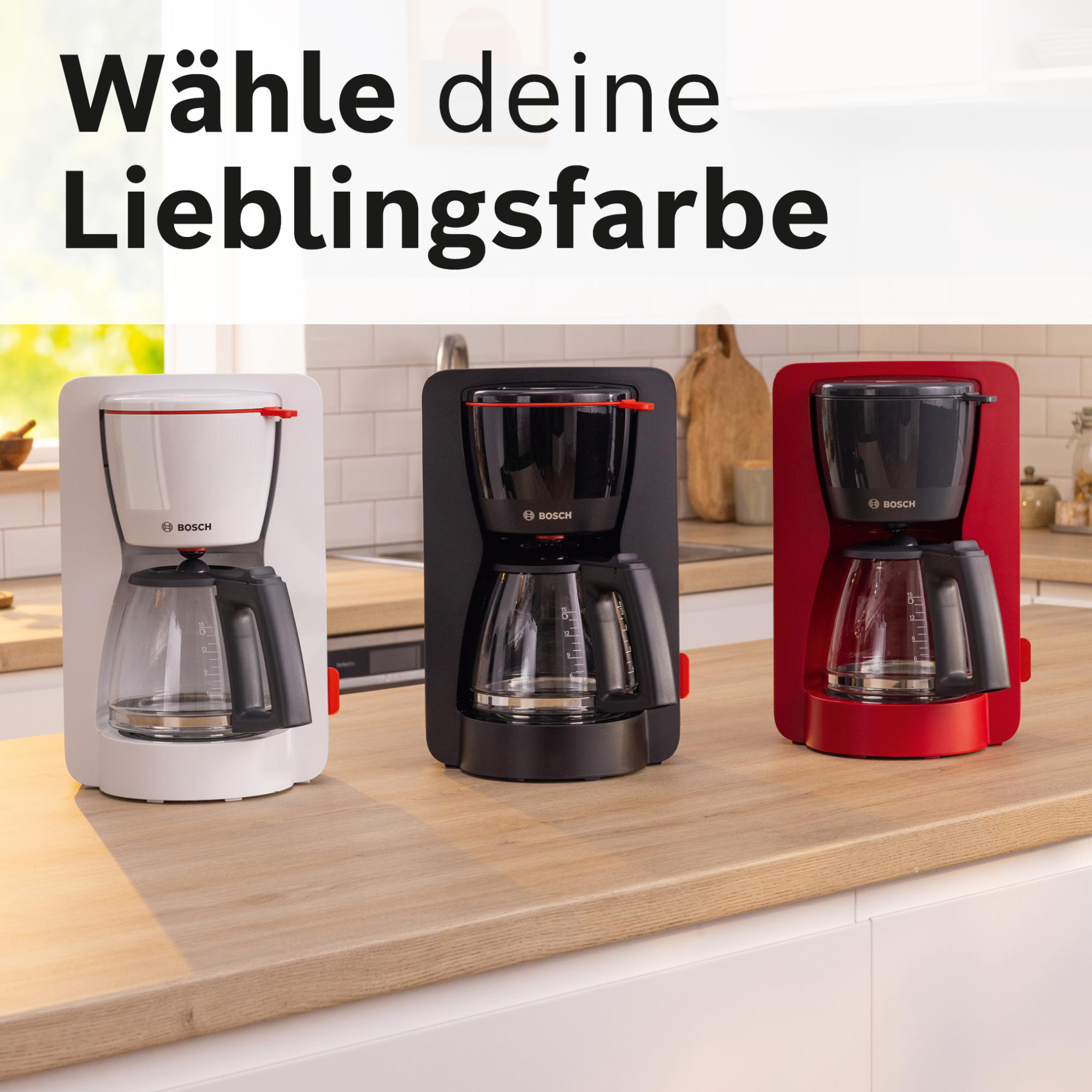 Drei Kaffeemaschinen: weiß, schwarz und rot. Sie stehen auf einer Holzoberfläche. Der Hintergrund ist eine Küche.