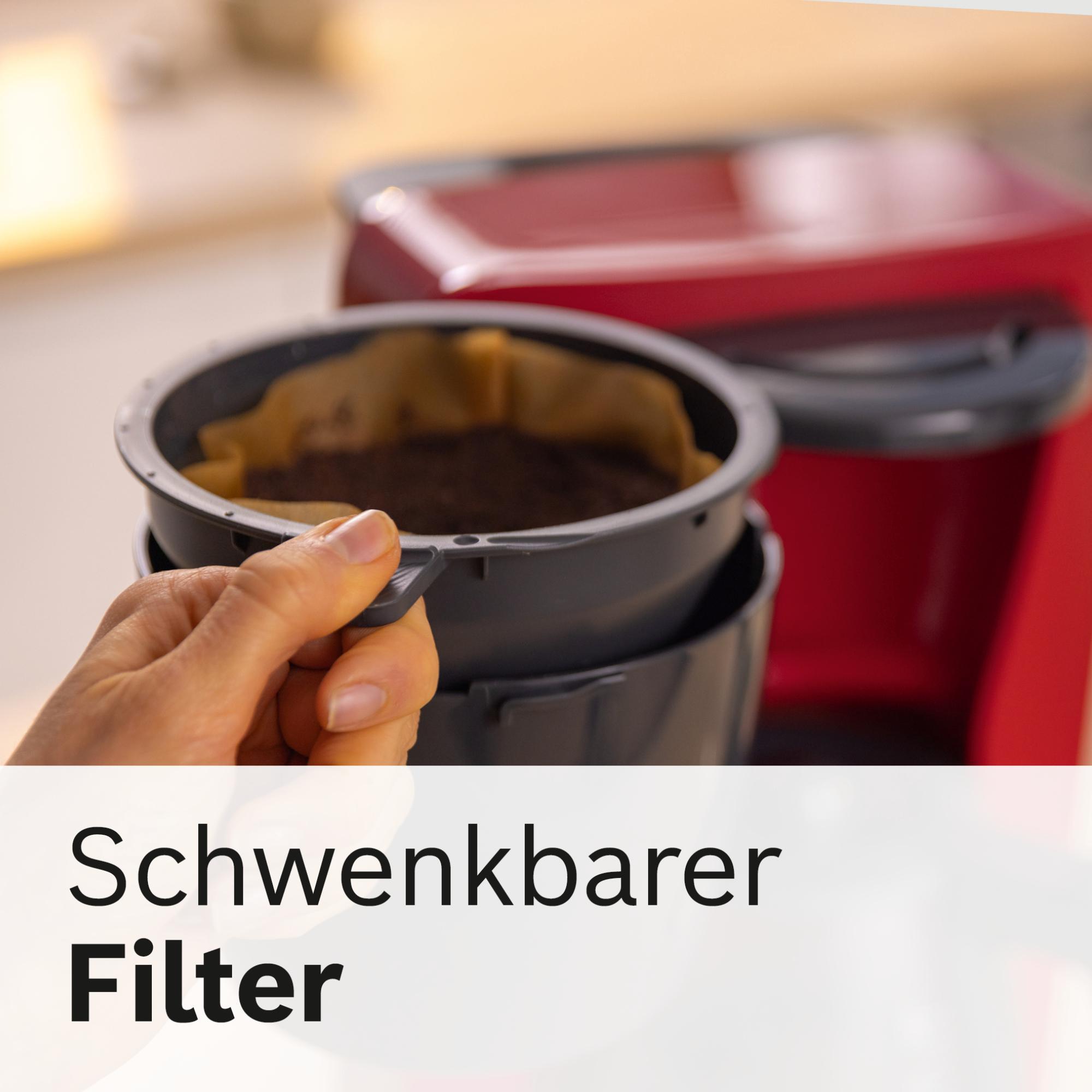 Eine Hand hält einen Kaffeefilter mit Kaffeepulver. Im verschwommenen Hintergrund befindet sich eine rote Kaffeemaschine.