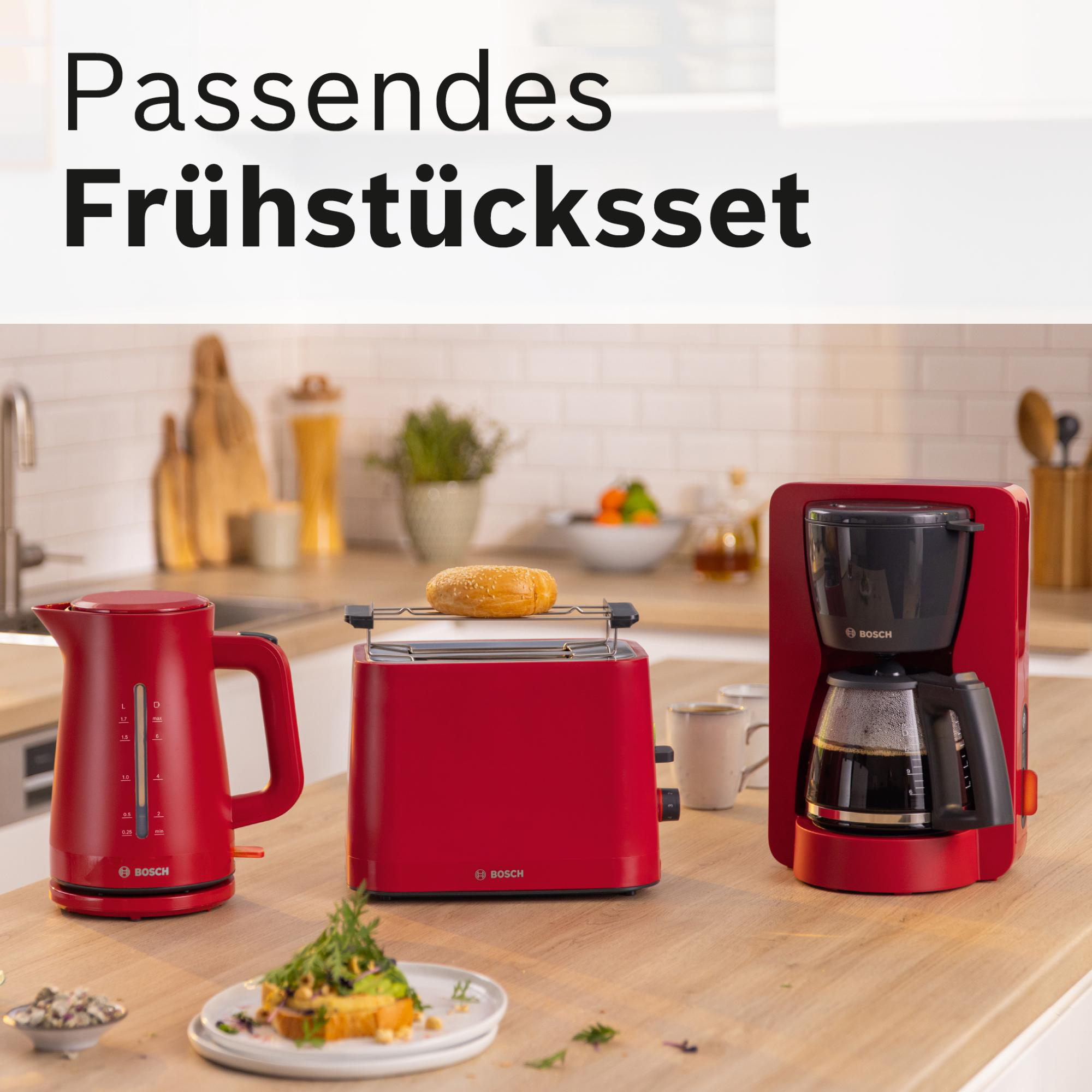 Ein roter Wasserkocher, Toaster und Kaffeemaschine auf einer Holztheke. Ein getoastetes Brötchen liegt im Toaster. Ein Teller mit Essen davor.