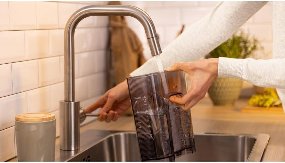 Person füllt einen transparenten Wasserbehälter aus einem silbernen Wasserhahn in einem Küchenbecken. Ein grauer Becher und eine Pflanze sind ebenfalls sichtbar.