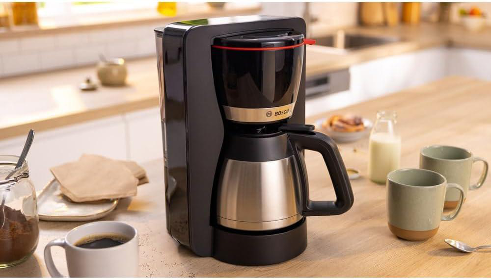 Ein schwarzer Bosch-Kaffeeautomaten steht auf einer Holzoberfläche. Daneben stehen Kaffeetassen, eine Milchflasche und Kaffeepulver.
