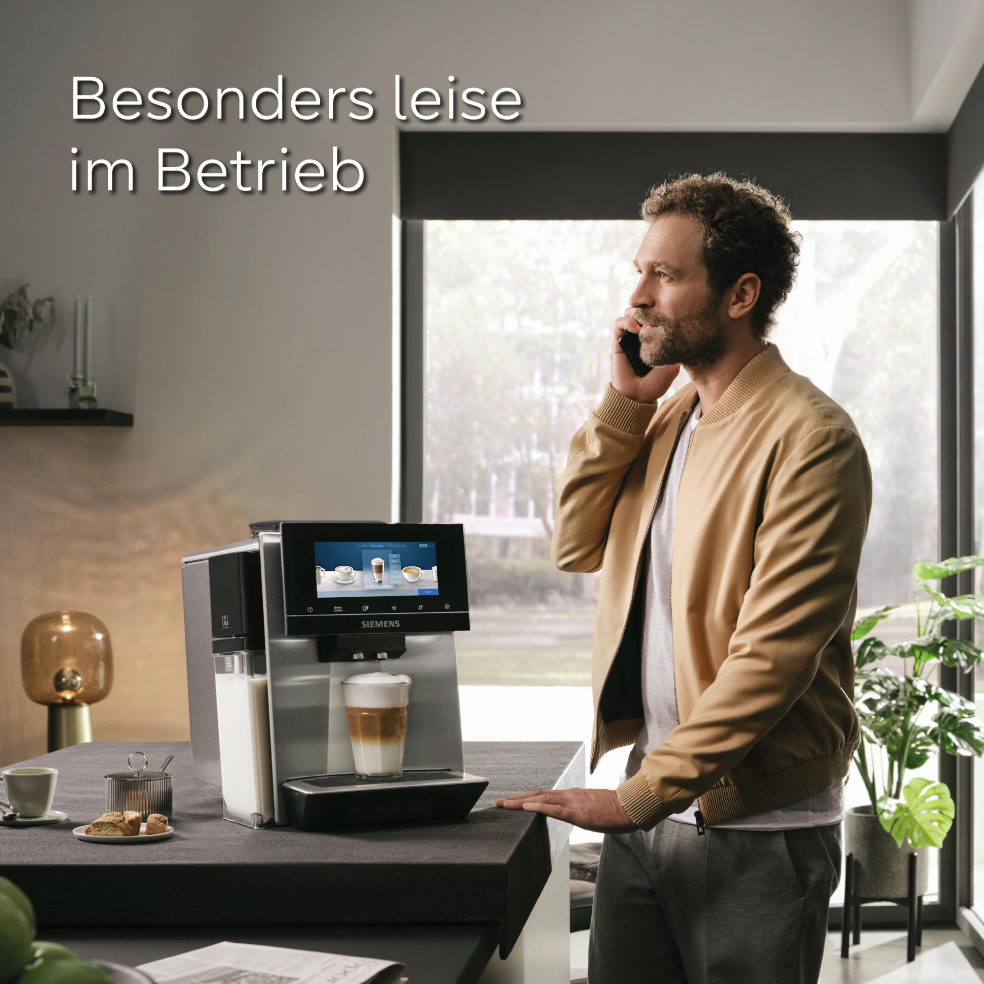 Mann telefoniert neben Kaffeemaschine. Latte im Glas. Text 'Besonders leise im Betrieb'. Moderner Küchenhintergrund.