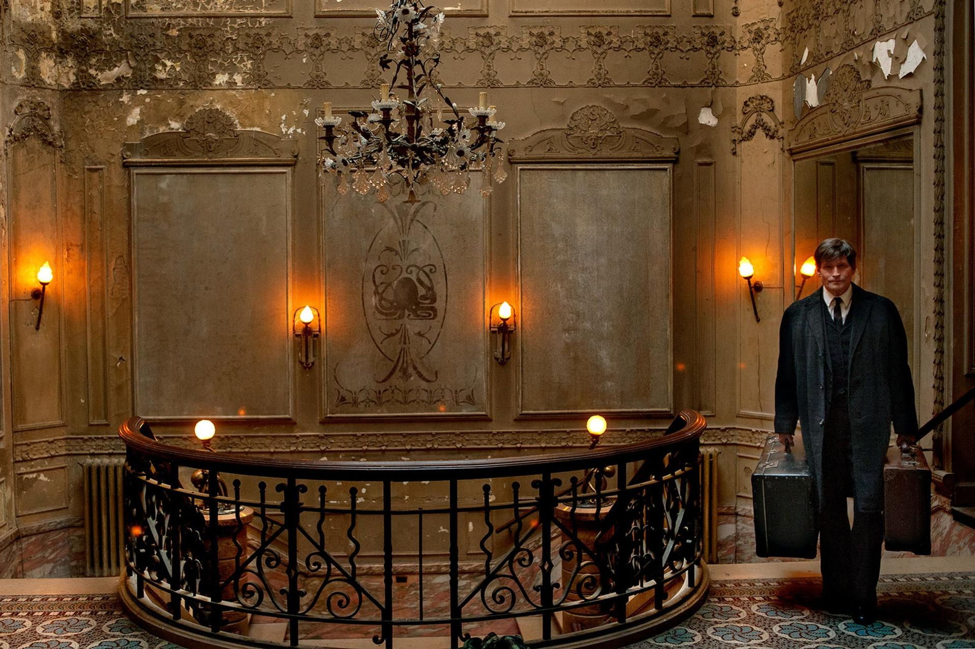 Ein Mann in einem dunklen Mantel mit Koffern steht in einer prächtigen, verzierten Halle mit Kronleuchter und Ziergeländer.