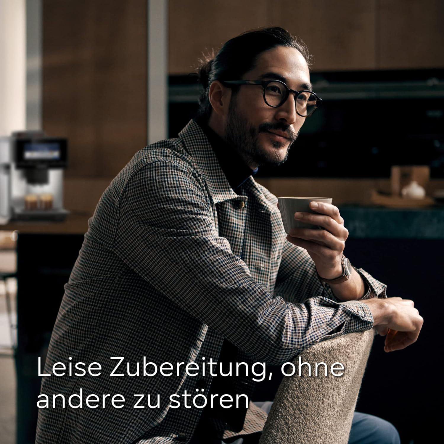 Mann mit Brille hält eine Tasse und sitzt. Kaffeemaschine im Hintergrund. Er trägt eine karierte Jacke, dunkle Haare.