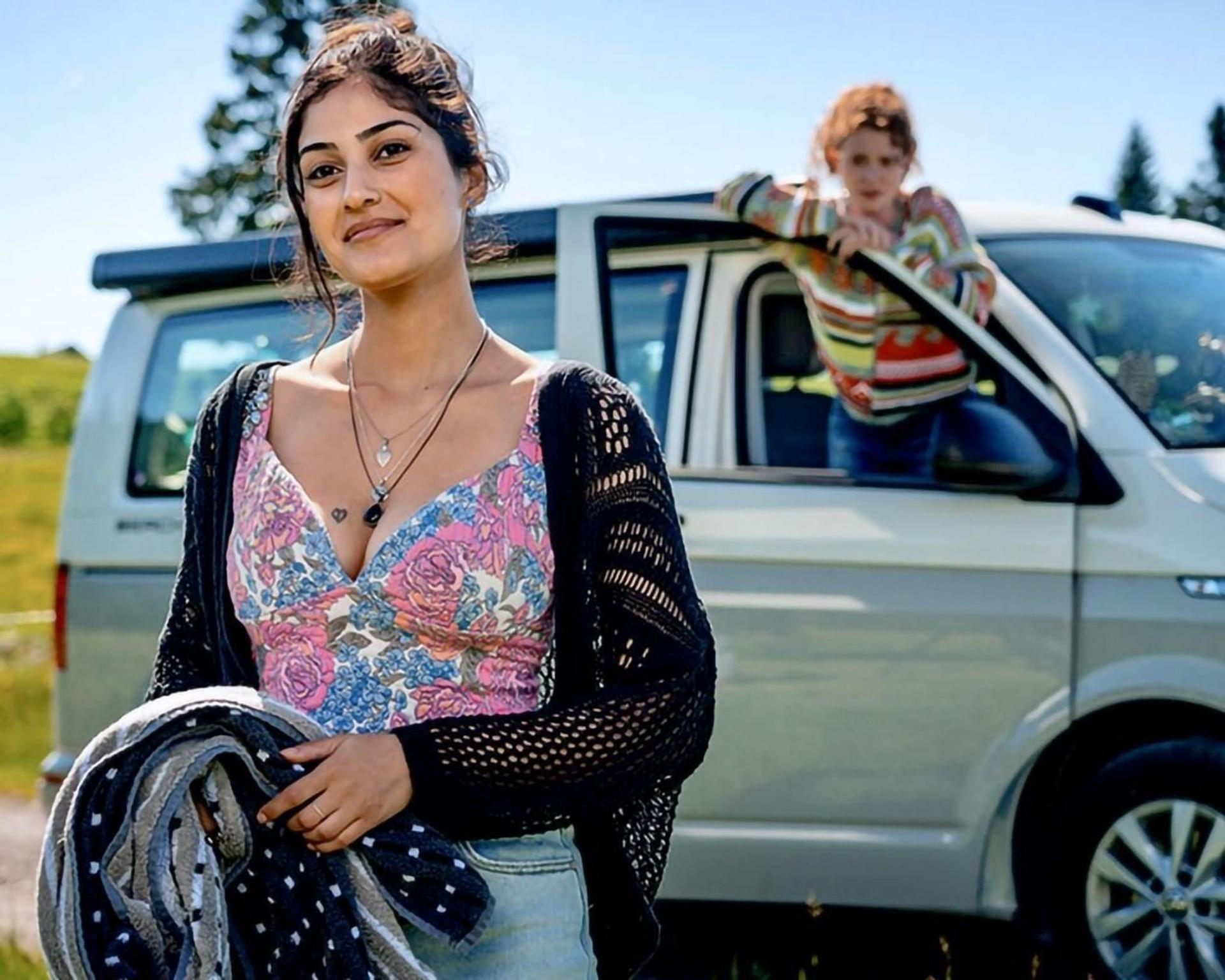 Eine Frau mit floralem Oberteil und schwarzer Strickjacke lächelt. Ein Kind lehnt sich aus einem Van-Fenster. Feld und blauer Himmel im Hintergrund.