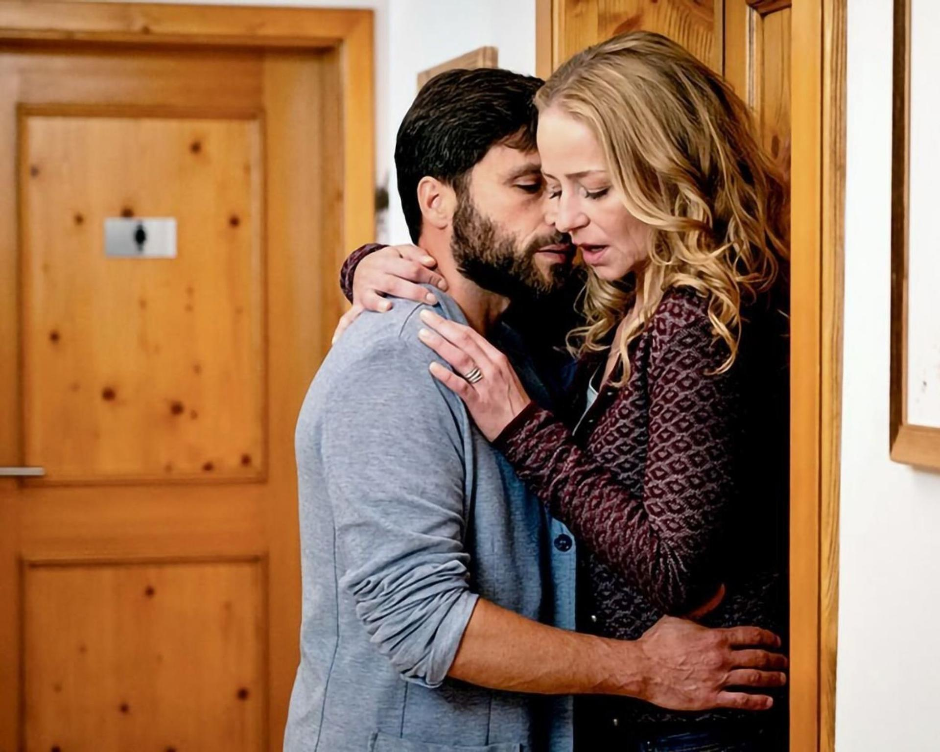 Ein Mann mit Bart umarmt eine Frau. Sie stehen in der Nähe einer Holztür. Die Frau hat blondes Haar und trägt eine gemusterte Jacke.