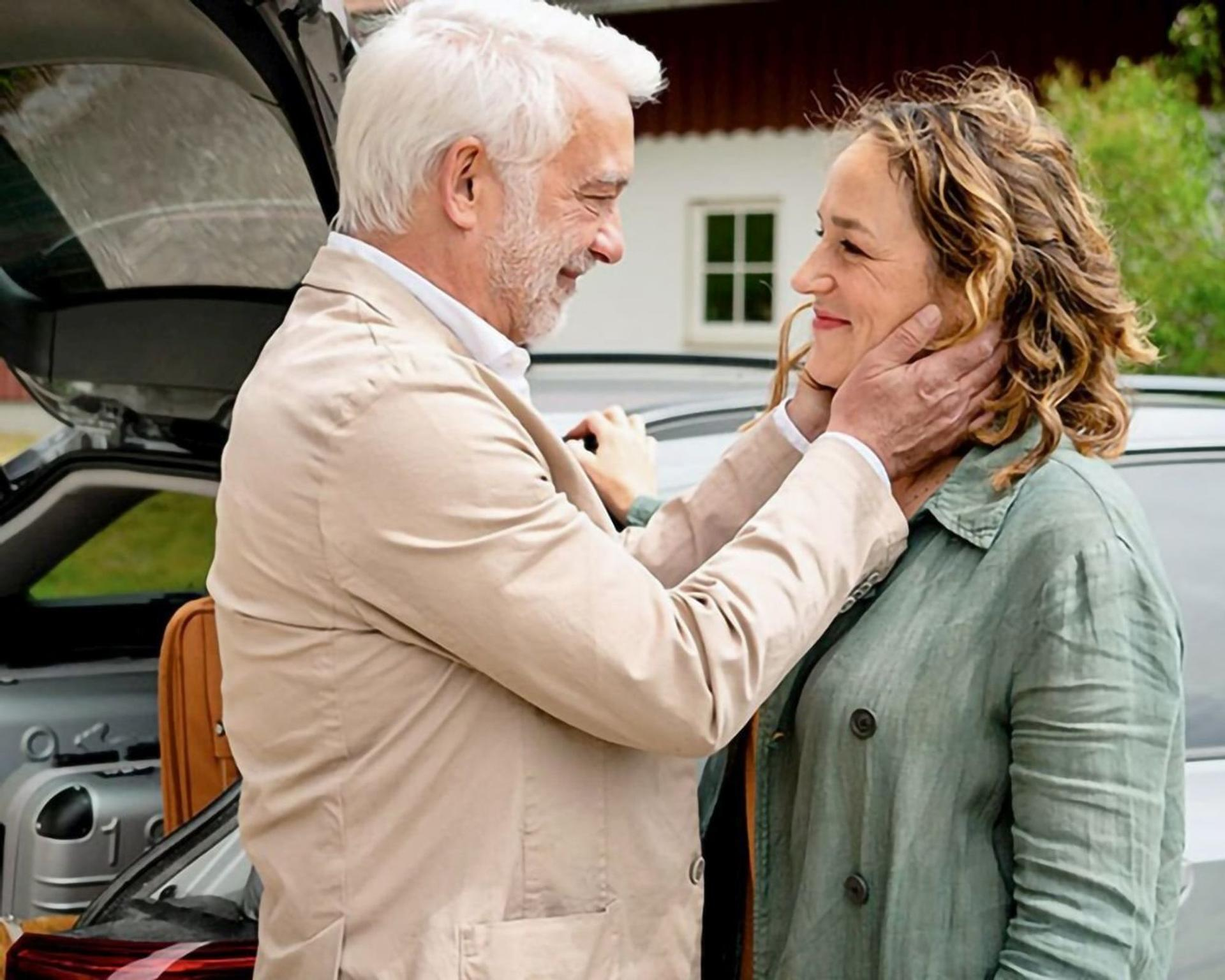Ein älterer Mann hält zärtlich das Gesicht einer Frau. Sie stehen an einem offenen Kofferraum. Im Hintergrund ein Gebäude mit Fenster.