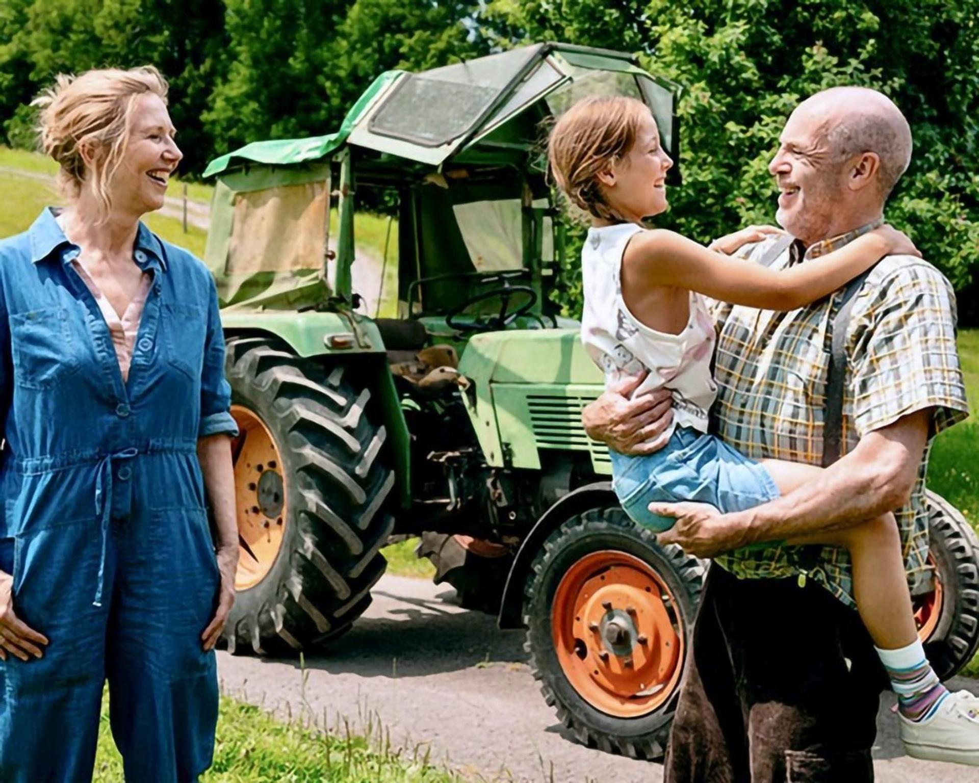 Ein Mann hält ein Kind vor einem grünen Traktor, eine Frau steht daneben.