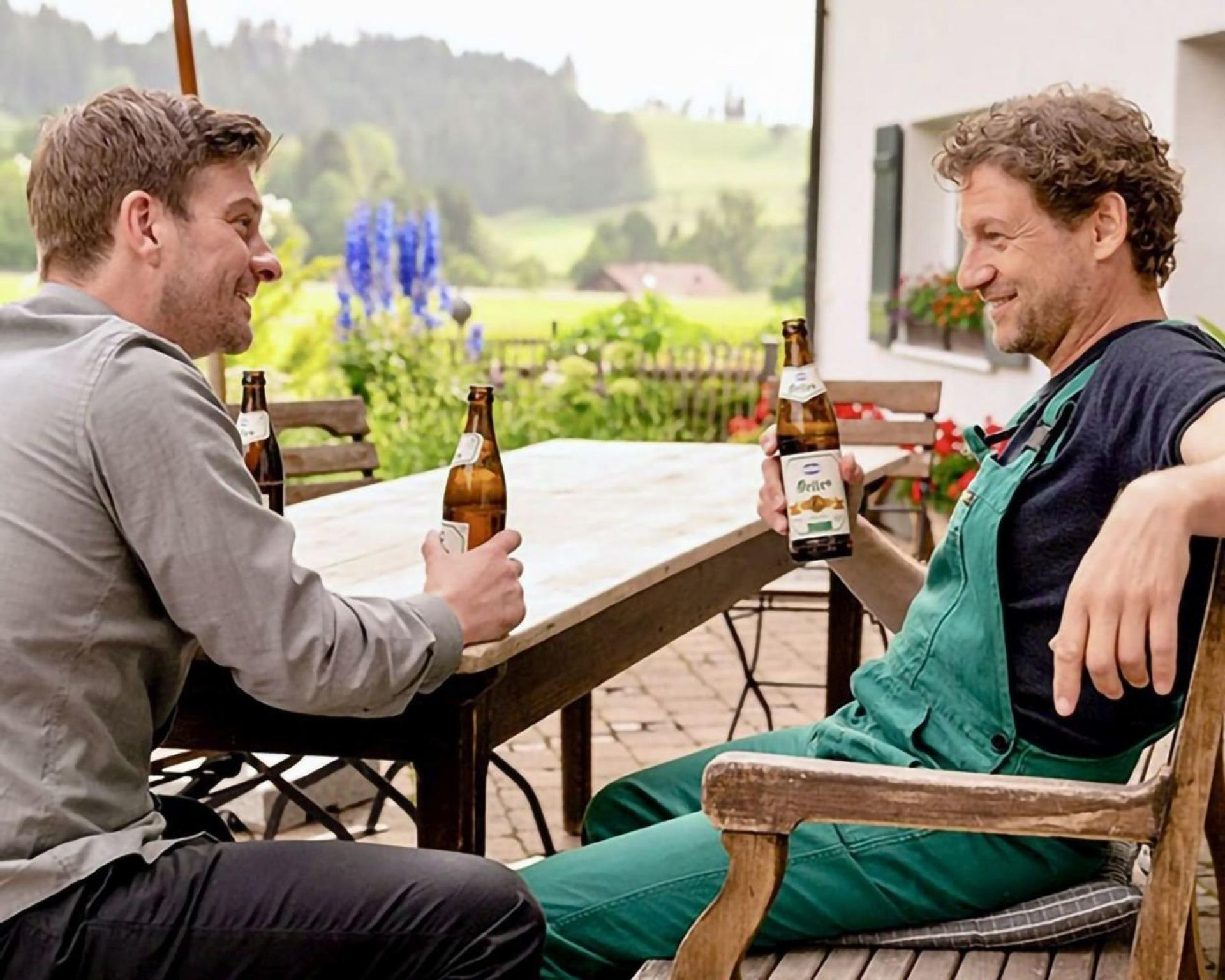 Zwei Männer sitzen an einem Tisch und trinken Bier. Im Hintergrund ist eine Landschaft.