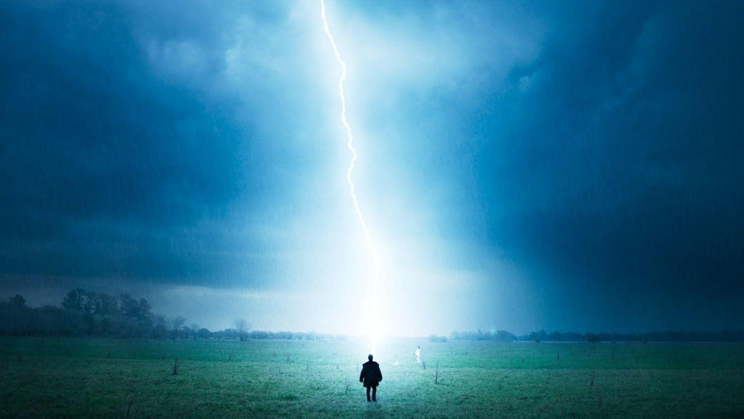 Eine Person steht auf einem Feld, während ein Blitz einschlägt. Dunkelblauer Himmel. Eine zweite Person ist im Hintergrund. Bäume und Gras im Feld.