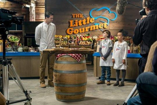 Ein Koch steht vor zwei Kindern in einer Küche mit dem Logo „The Little Gourmet“.
