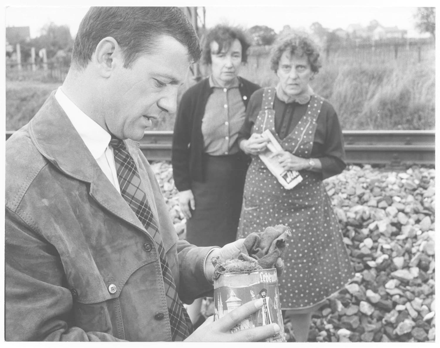 Mann im Mantel betrachtet eine Dose, zwei Frauen hinter ihm auf Bahngleisen. Felsen und Vegetation im Hintergrund.