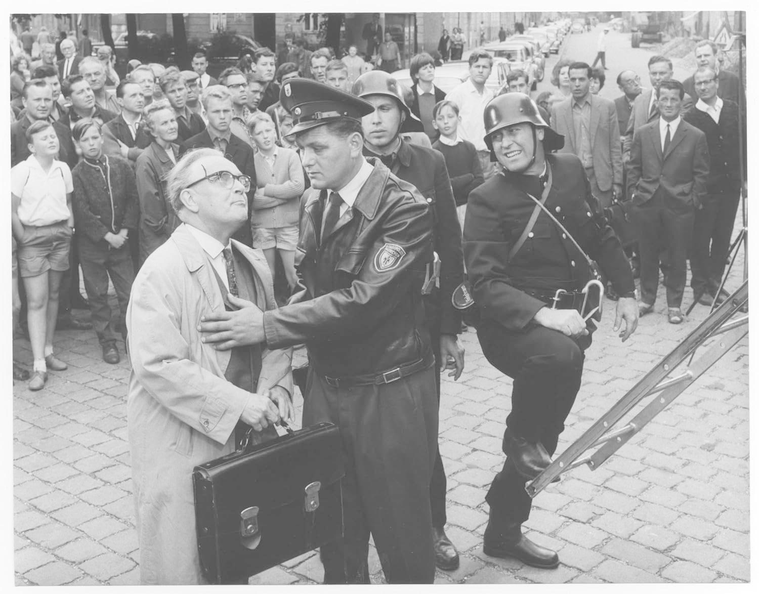 Eine Gruppe von Menschen, darunter uniformierte Beamte, umringt einen Mann im Anzug. Eine Leiter ist im Bild. Die Szene findet im Freien statt.