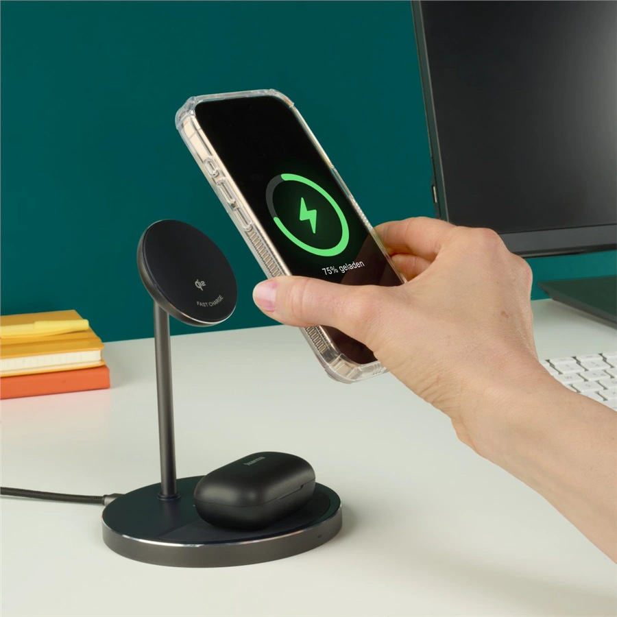 Ręka trzyma telefon ładujący się bezprzewodowo. Stacja ładująca ze stojakiem i słuchawkami na białym biurku. Zielone tło.