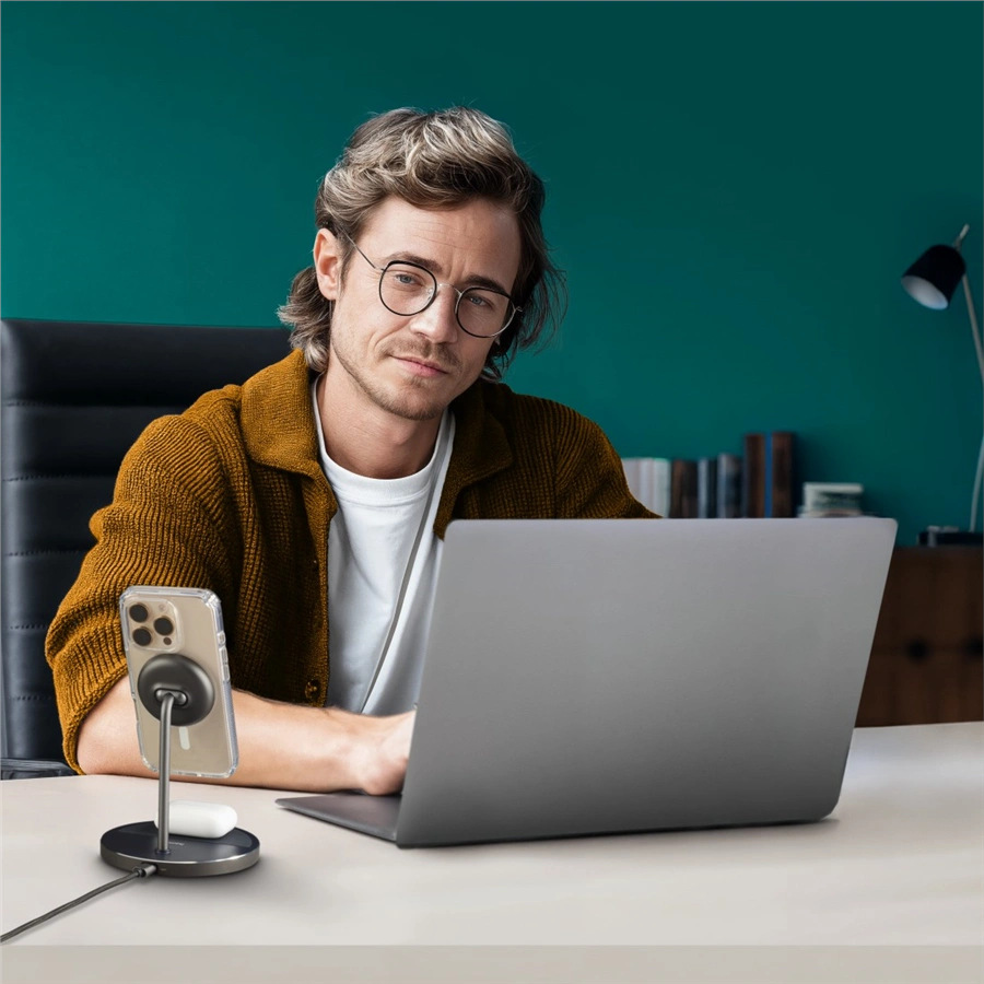 Mężczyzna w okularach siedzi przy biurku, używając laptopa. Telefon i słuchawki znajdują się na stojaku do ładowania. Tło jest turkusowe.