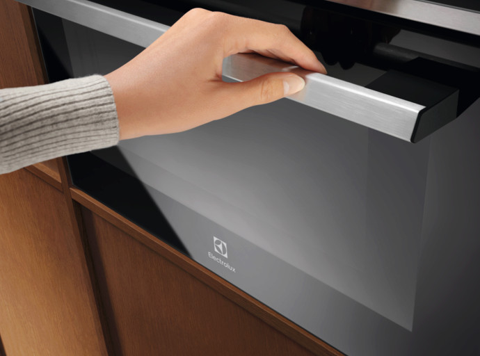 Ręka pociąga za uchwyt czarnego piekarnika. Piekarnik znajduje się w drewnianej szafce. Piekarnik ma logo Electrolux.