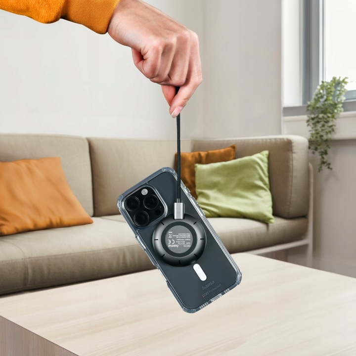 Dłoń trzyma telefon z kablem do ładowania. W tle sofa i stół. Telefon ma przezroczyste etui.