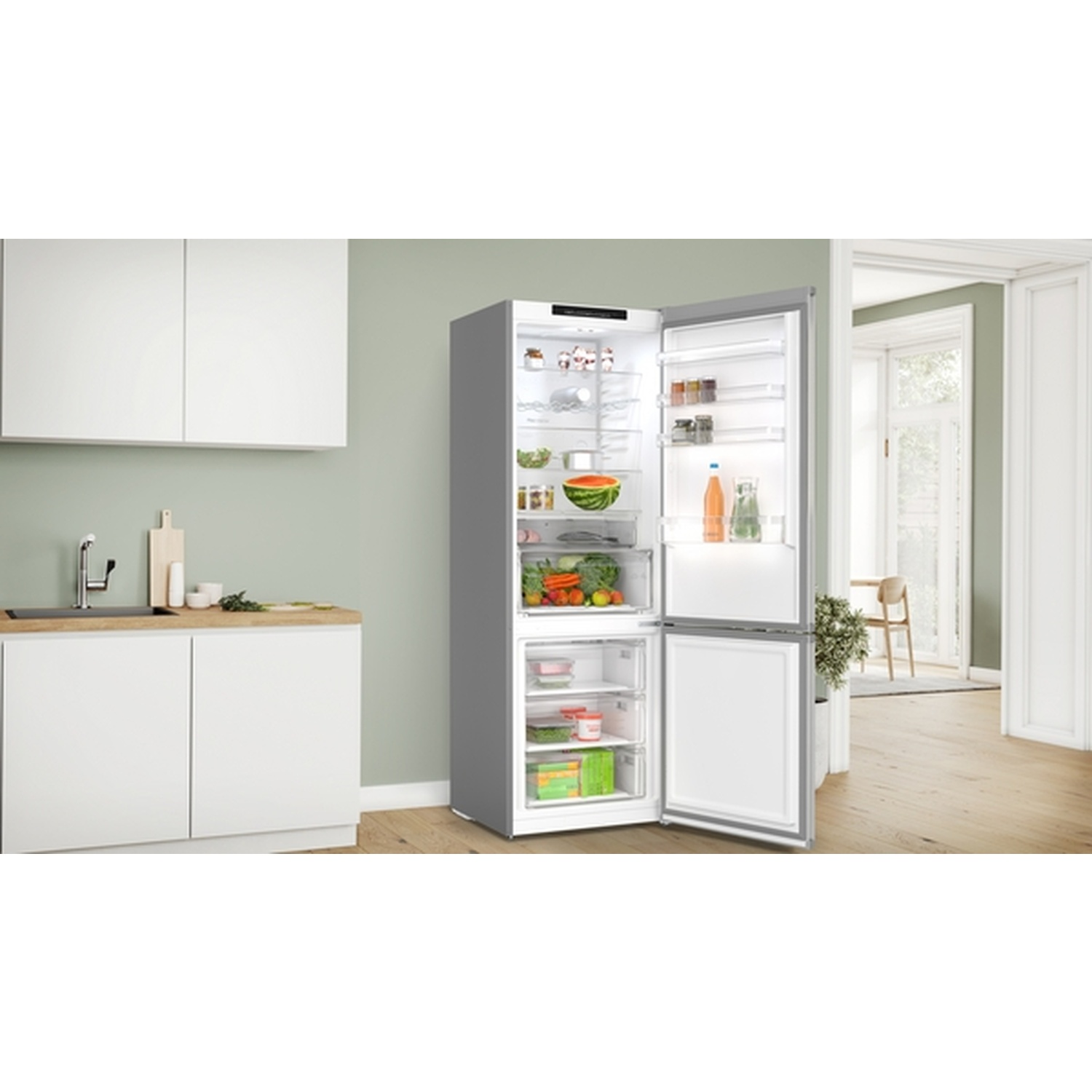 An open gray refrigerator in a kitchen. Inside, fruits, vegetables, and drinks are visible. A counter and a window are in the background.