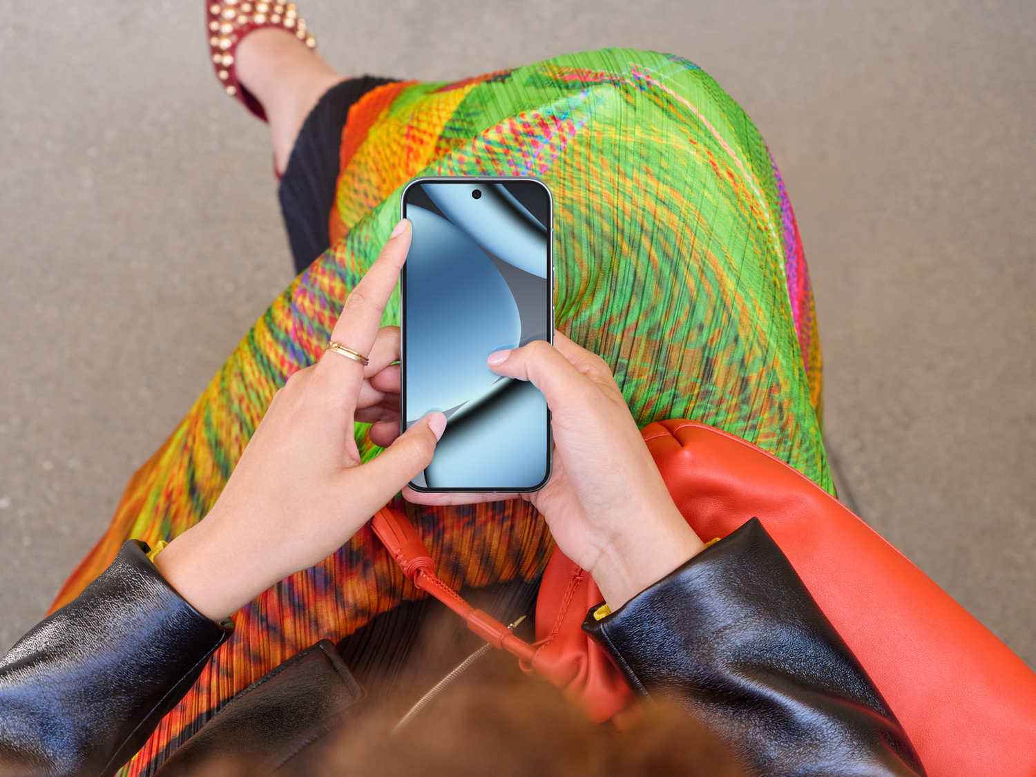 Una persona sostiene un teléfono. Lleva una falda colorida, un bolso rojo, una chaqueta negra y zapatos rojos.