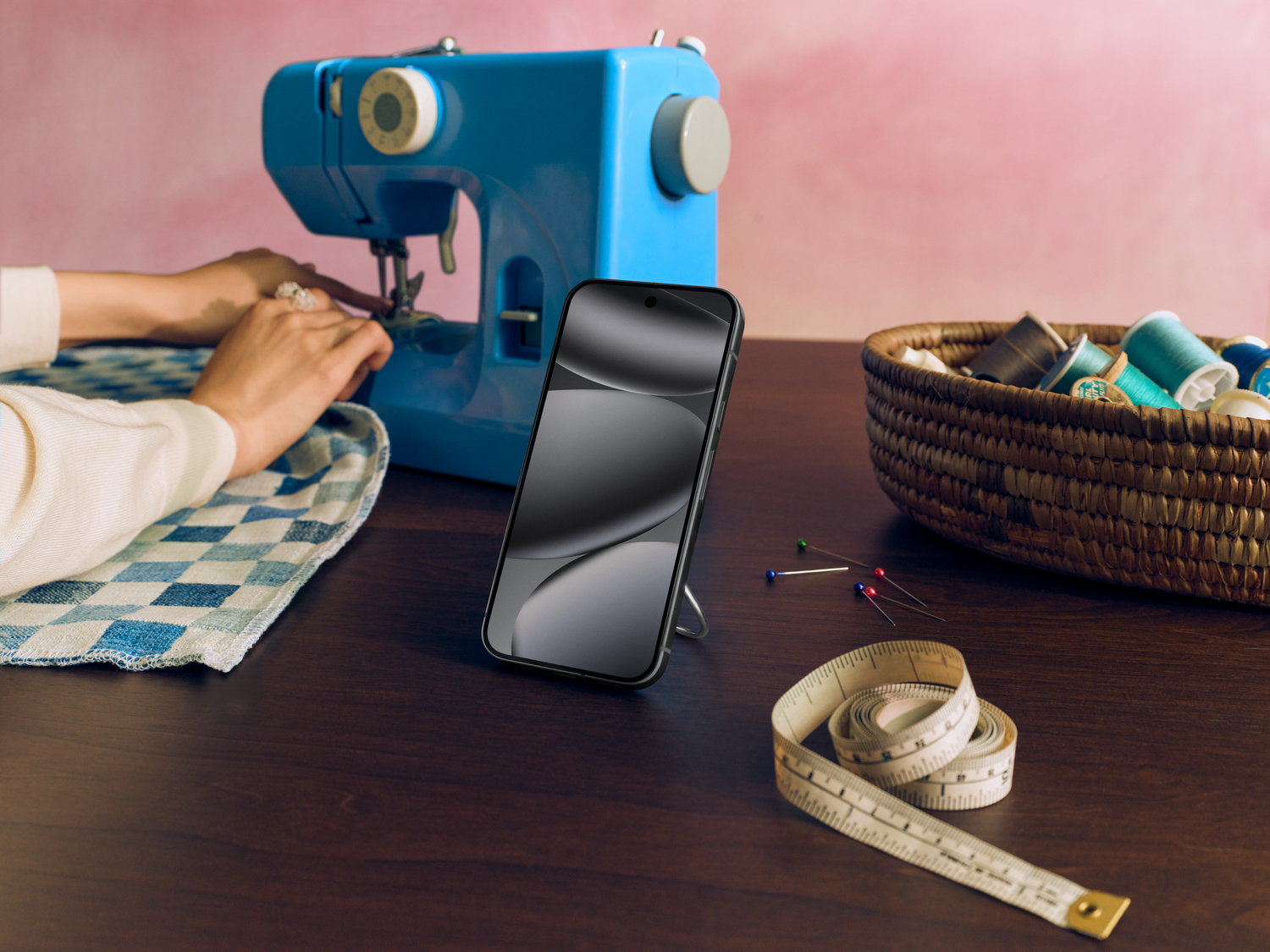 Una escena de costura muestra una máquina de coser azul, un teléfono y una cesta con carretes de hilo. Manos trabajan en la máquina; una cinta métrica es visible.
