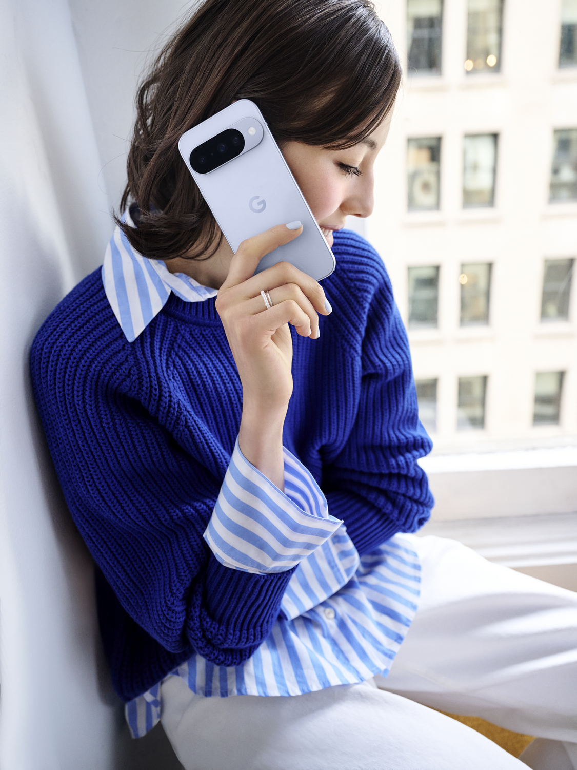 Una mujer sostiene un teléfono lavanda. Lleva un suéter azul sobre una camisa a rayas, pantalones blancos. Edificios de la ciudad en el fondo.