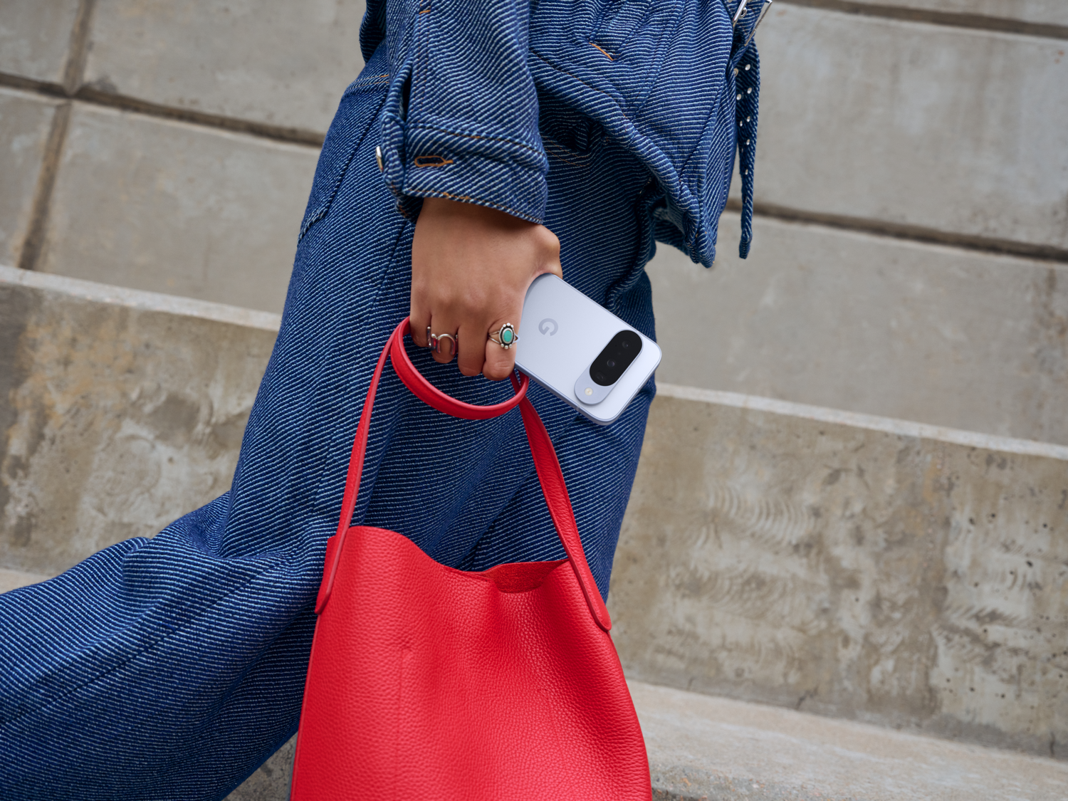 Persona con atuendo de mezclilla sosteniendo un teléfono y una bolsa roja, caminando por escaleras. Anillos en los dedos.