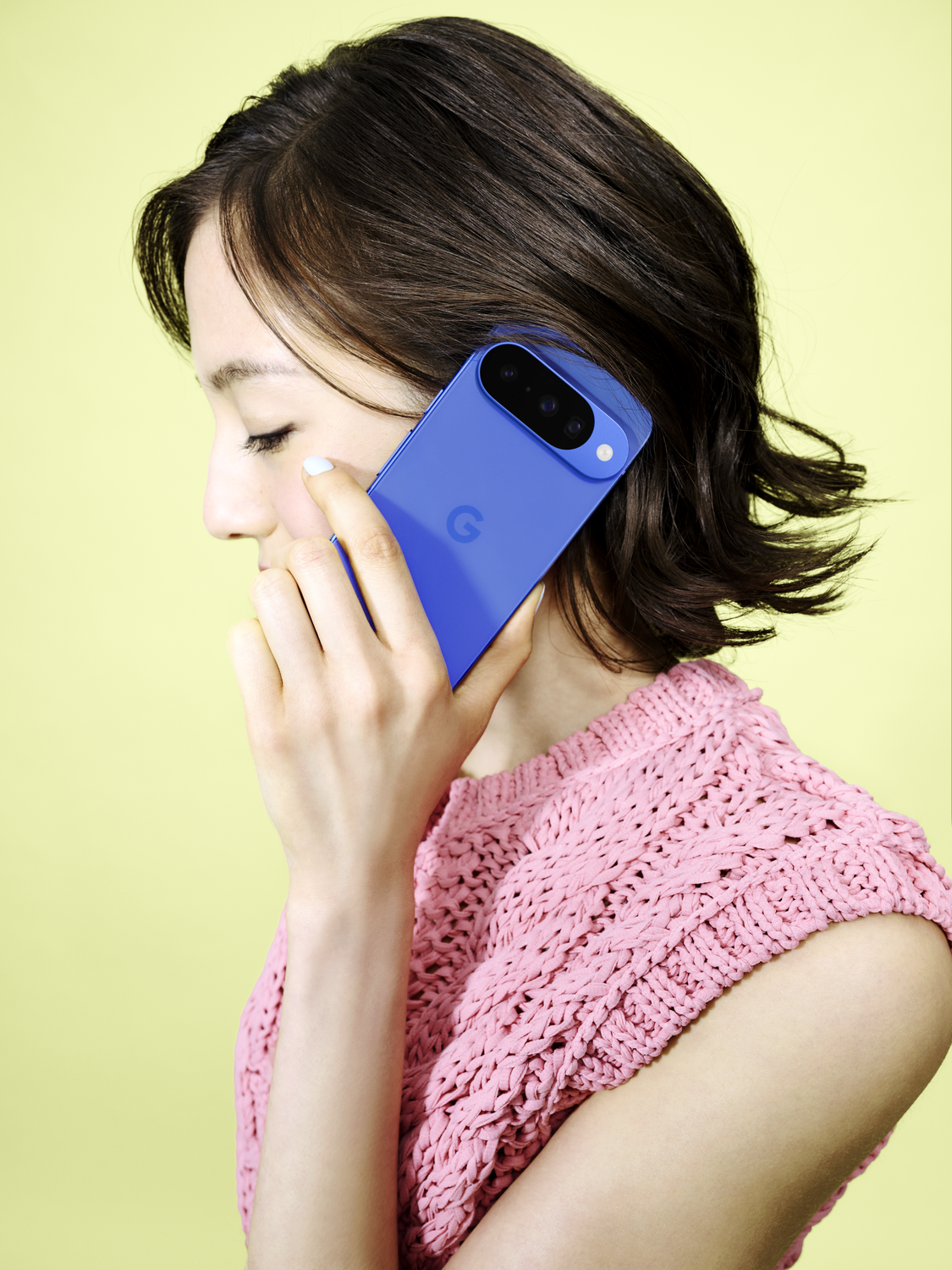 Mujer sosteniendo un teléfono azul en su oído. Lleva un suéter de punto rosa. Fondo amarillo.