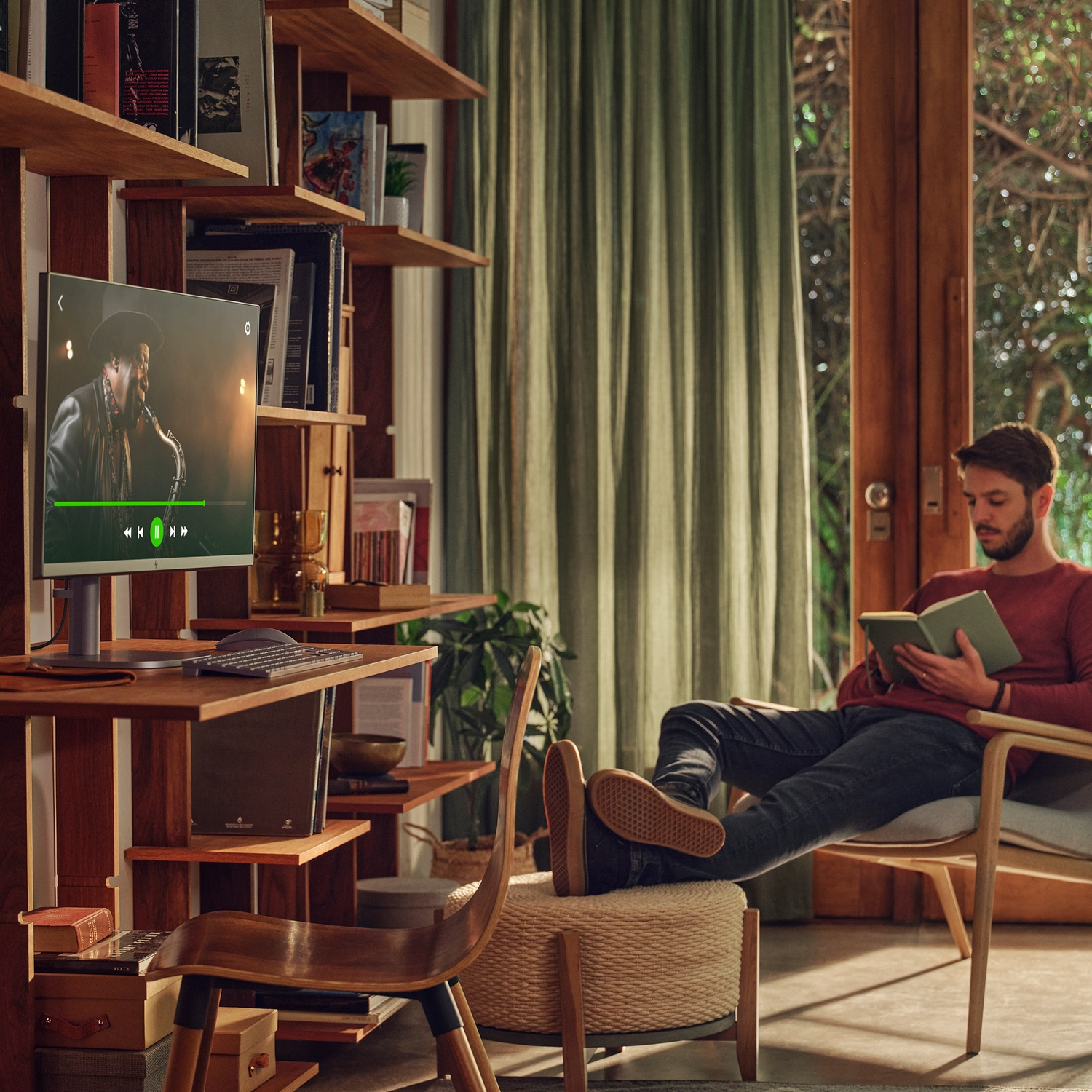 A man reads a book while another watches a video on a monitor. Bookshelves and a wooden chair are in the background.