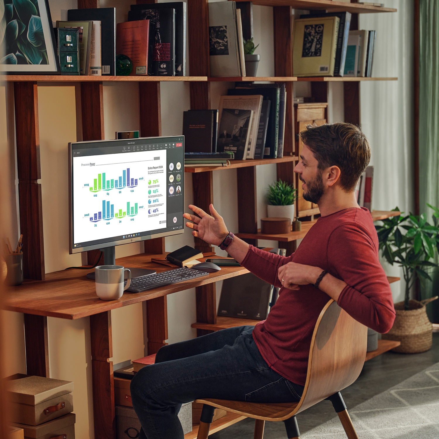 A man in a red shirt gestures at a computer screen displaying charts. He sits at a wooden desk with a keyboard, mug, and books in the background.