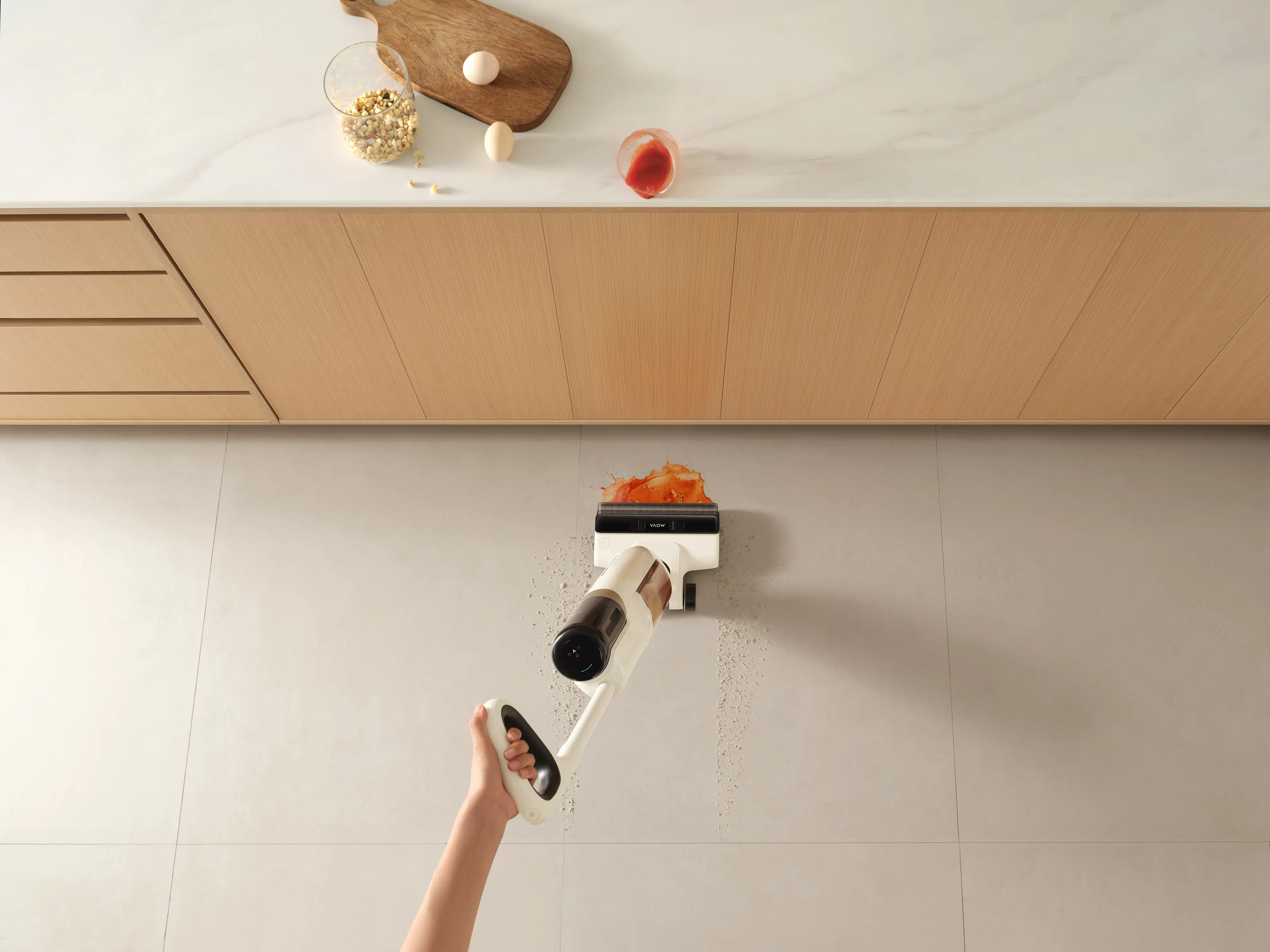 Ręka trzyma biały odkurzacz czyszczący rozbryzganą czerwoną substancję na jasnej podłodze. Produkty spożywcze na blacie.