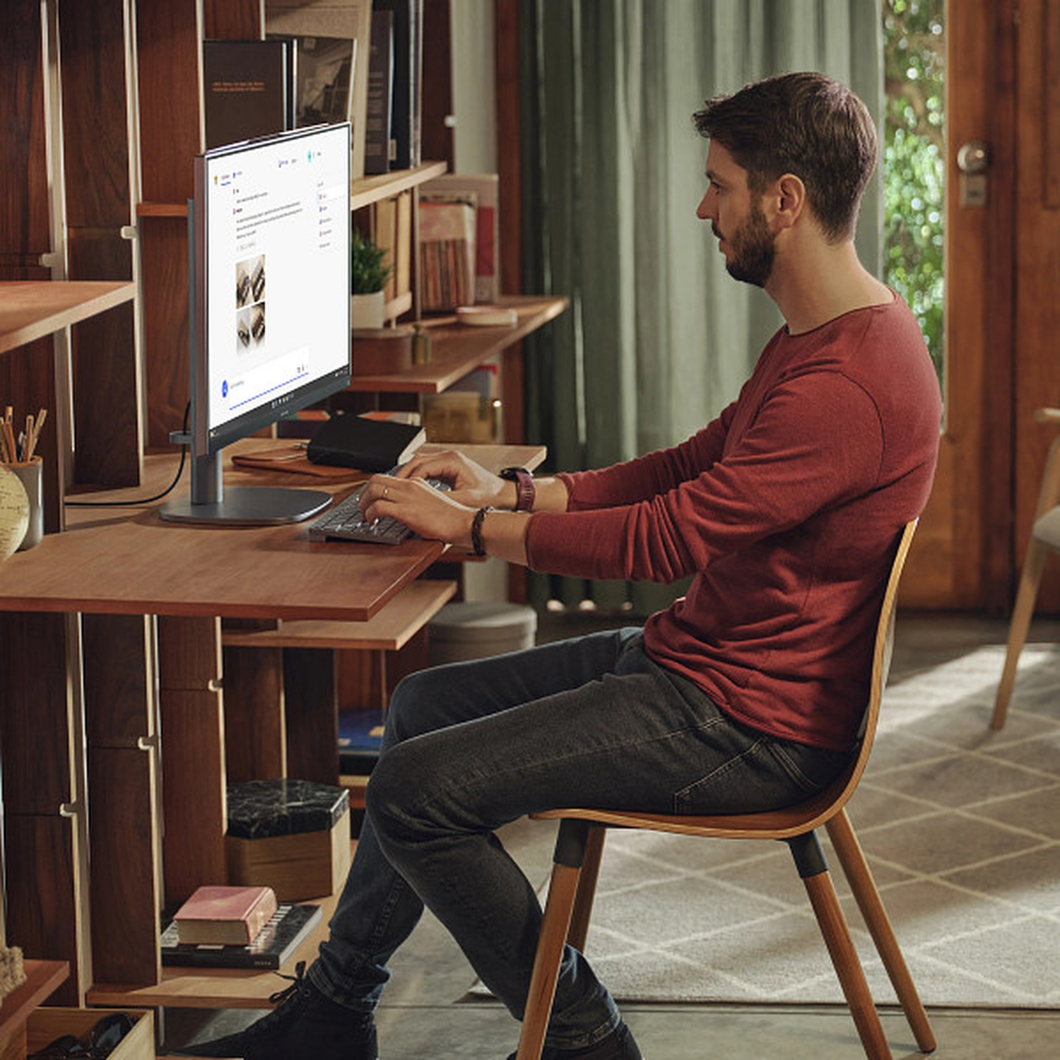 Homme en chemise rouge assis à un bureau, utilisant un ordinateur. Étagères en arrière-plan. Chaise et bureau en bois. Lumière naturelle.