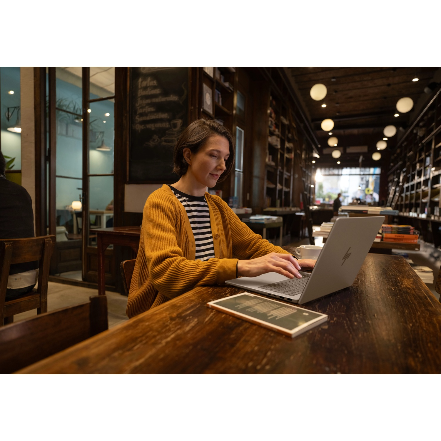 Vrouw in een café typt op een laptop. Ze draagt een gele vest en een gestreept shirt. Boekenkasten en een menu zijn zichtbaar.
