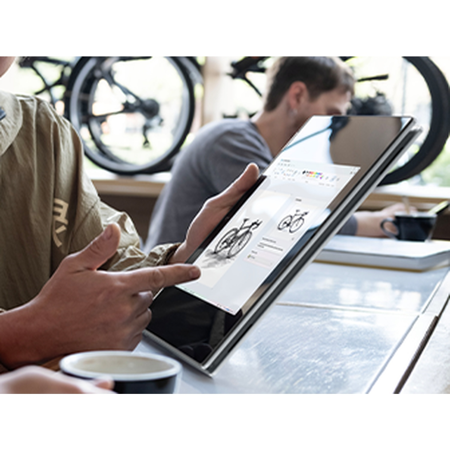 Person pointing at a tablet displaying bicycle designs. Another person in the background, and a coffee cup in the foreground.