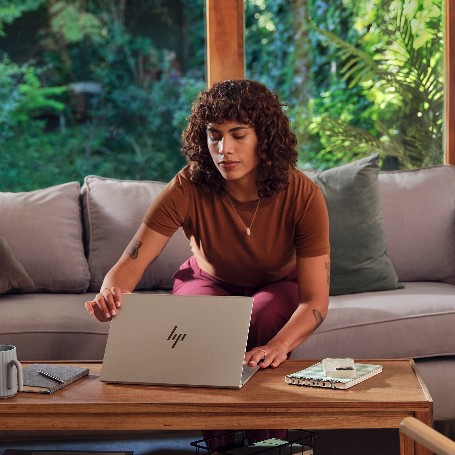 Een vrouw zit op een bank en opent een laptop op een houten tafel. Er is een notitieboekje, een mok en groen in de achtergrond.