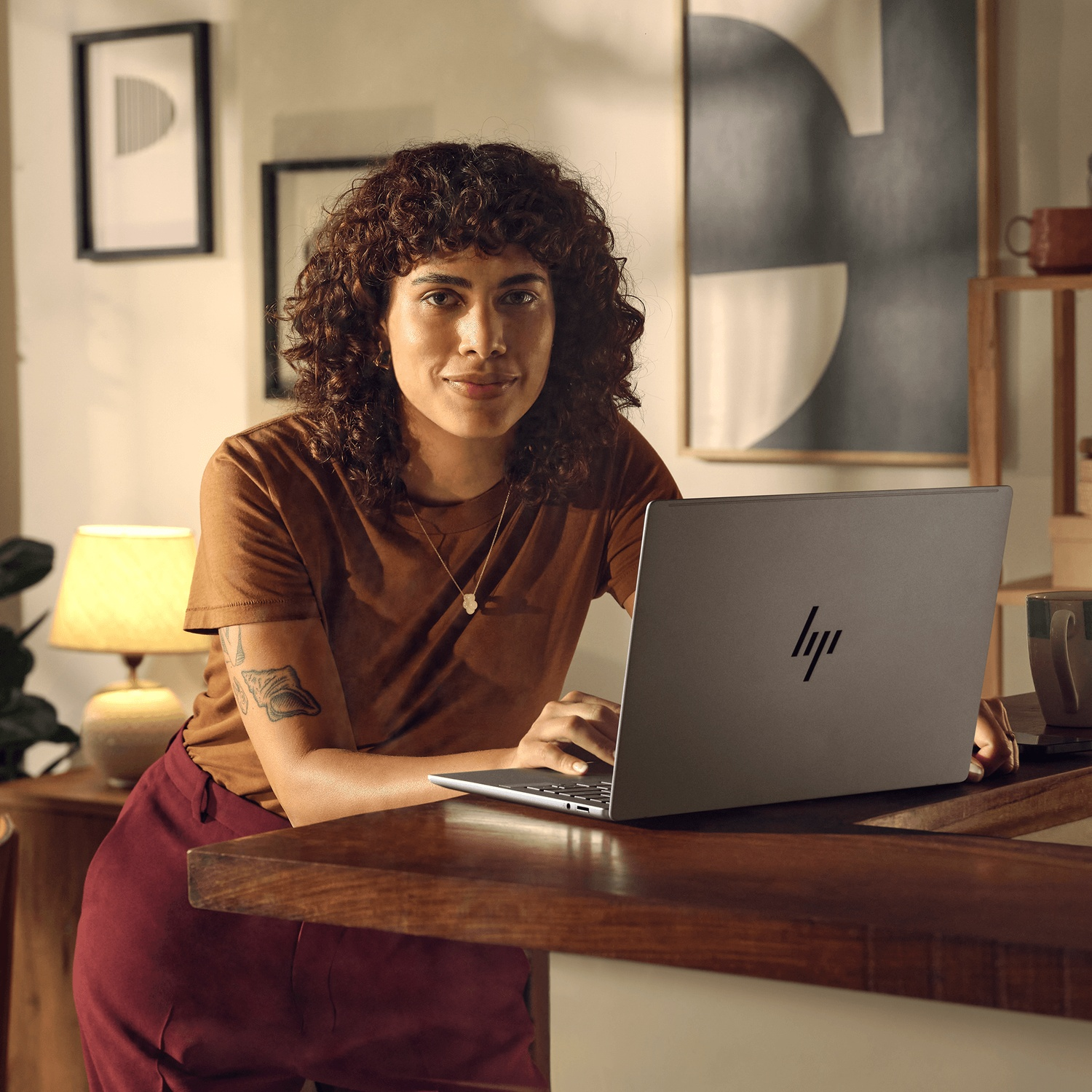 Een vrouw met krullend haar, bruine top, rode broek, gebruikt een laptop. Ze leunt op een houten oppervlak.