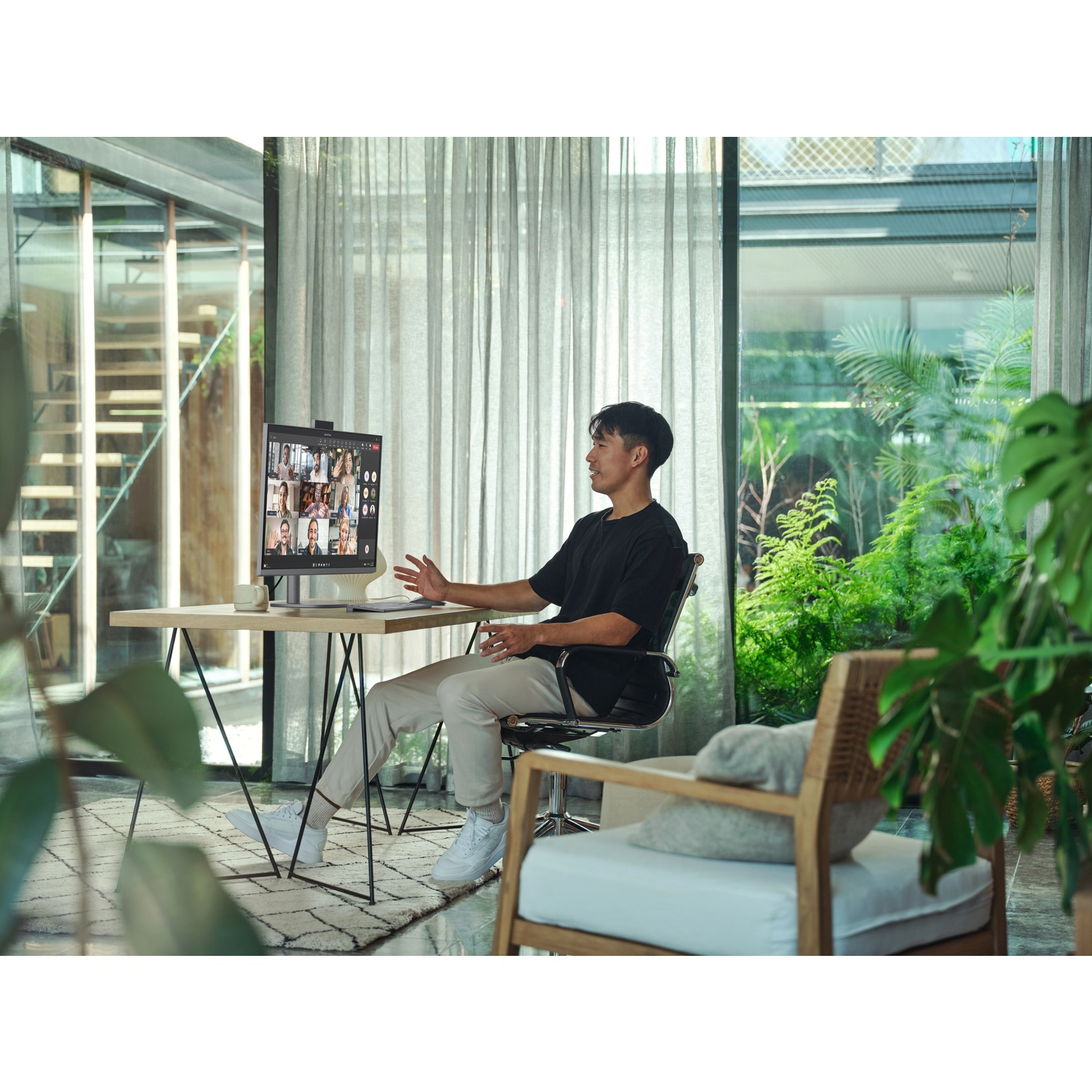 Een man in een zwart shirt is in een videogesprek. Een monitor toont andere deelnemers. Een tafel, stoel en planten omringen hem.