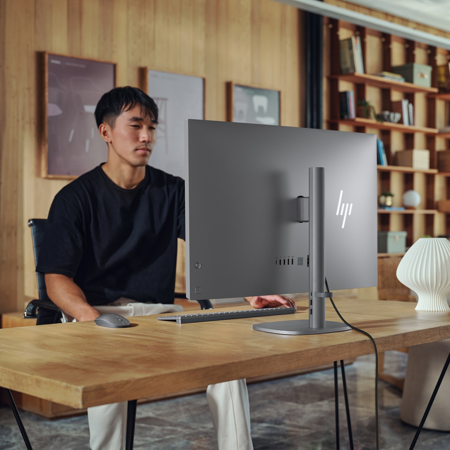 Een man zit aan een bureau met een monitor. Een toetsenbord en muis liggen voor hem. Schappen en kunstwerken op de achtergrond.