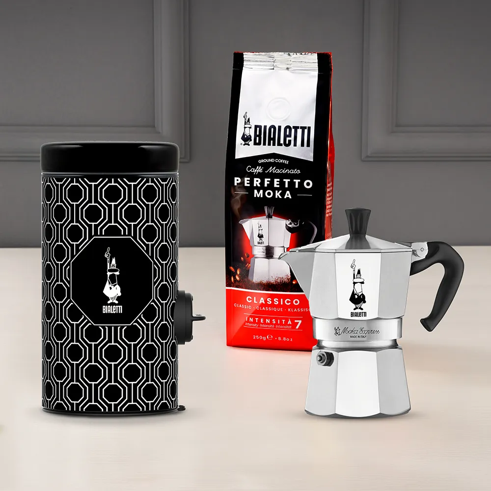 Kávészett: egy őrlő, egy kávézacskó és egy ezüst Moka kávéfőző. Mindegyik Bialetti márkájú, világos felületen elrendezve.