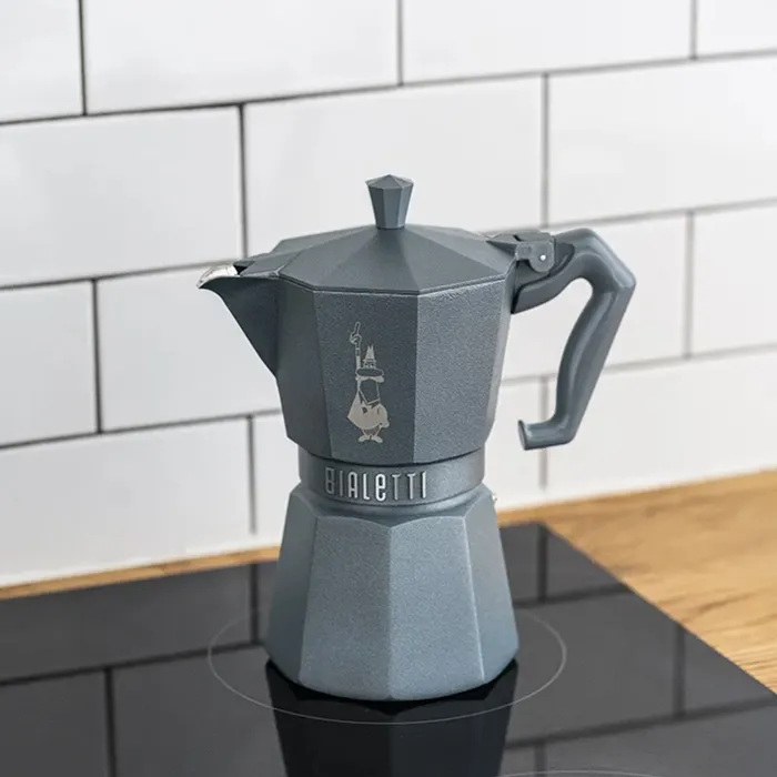 Egy szürke Bialetti kávéfőző egy fekete főzőlapon. Fehér csempézett fal a háttérben.