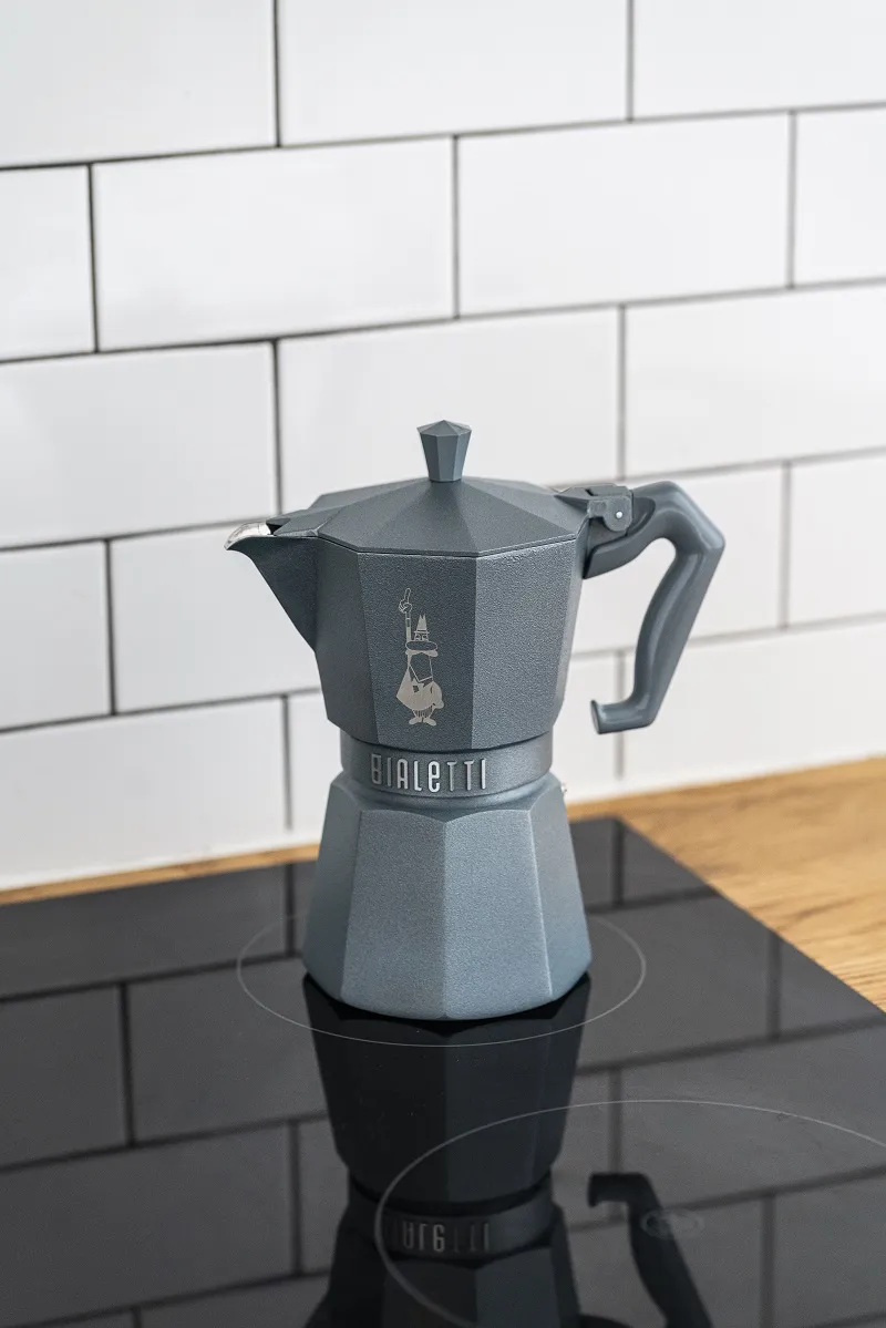 Egy szürke Bialetti mokka kávéfőző egy fekete főzőlapon, fehér csempézett fal és fa pult előtt.