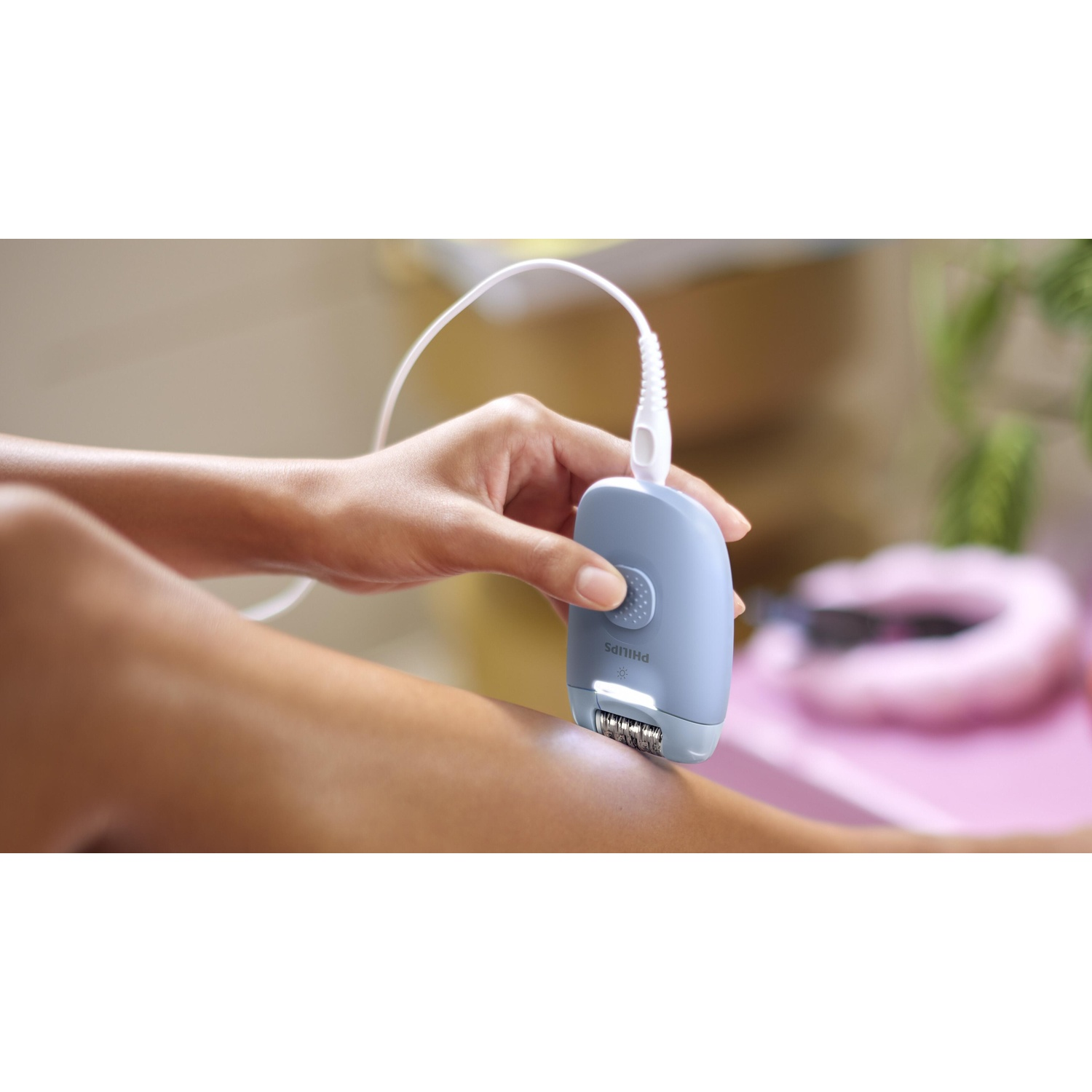 A person is using a blue epilator on their leg. The epilator has a white cord. The background is blurred with pink and green.