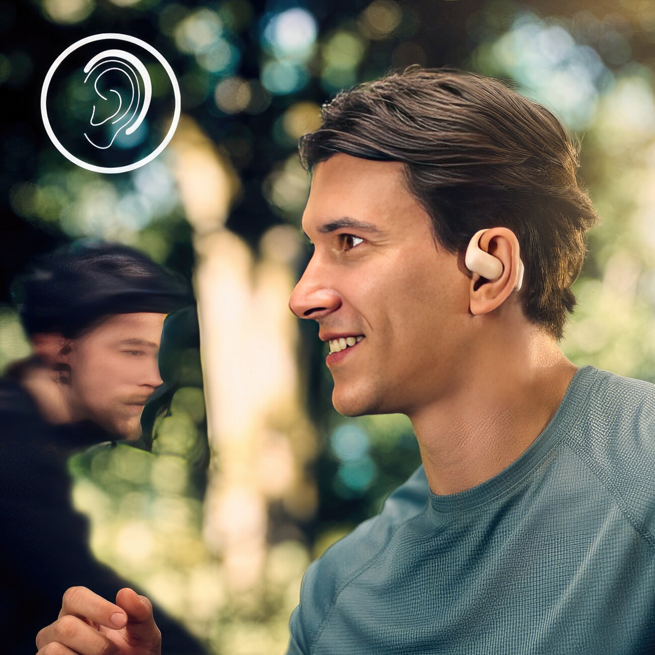 Mężczyzna uśmiecha się, nosząc aparat słuchowy. Rozmyta osoba i zielone liście w tle.