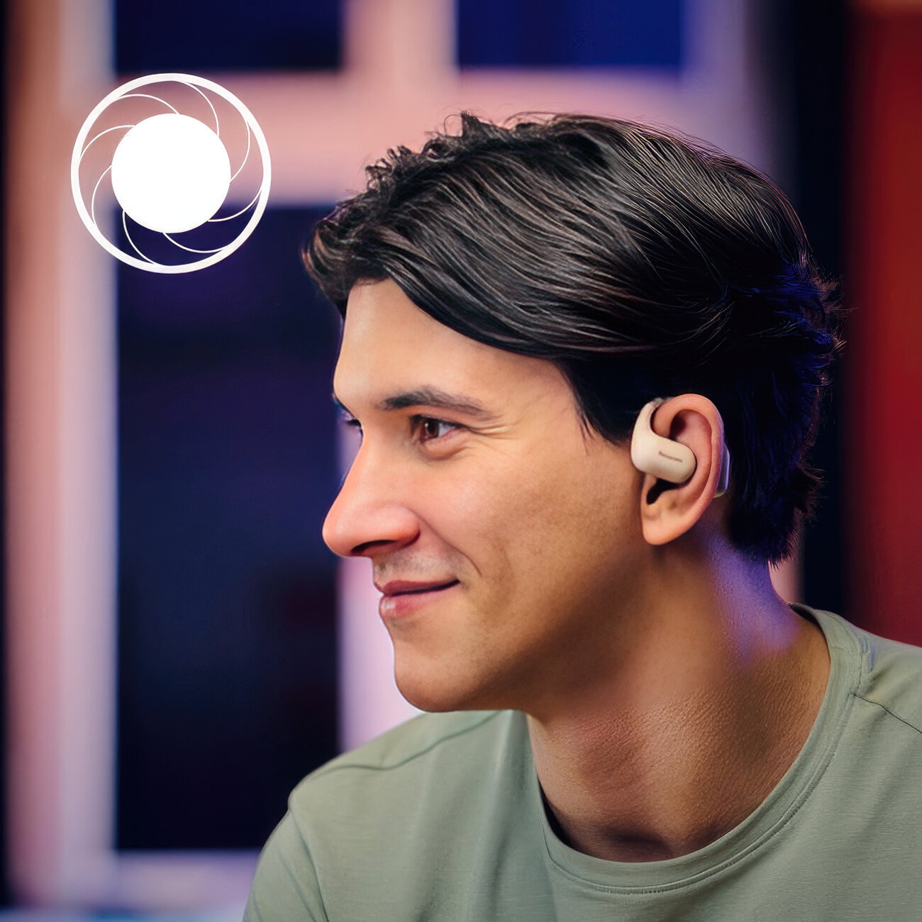 Mężczyzna noszący aparat słuchowy uśmiecha się. Ma ciemne włosy i zieloną koszulę. Tło jest rozmyte z niebieskim i czerwonym.